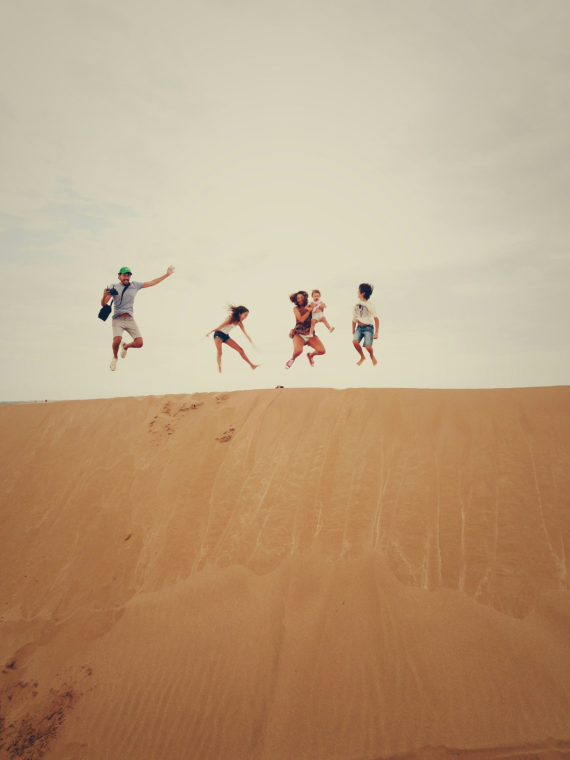 A family of five jumping on a sand dune in a desert, with the sky in the background.