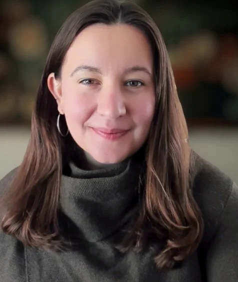 A woman with long brown hair wearing a gray turtleneck sweater smiling at the camera.