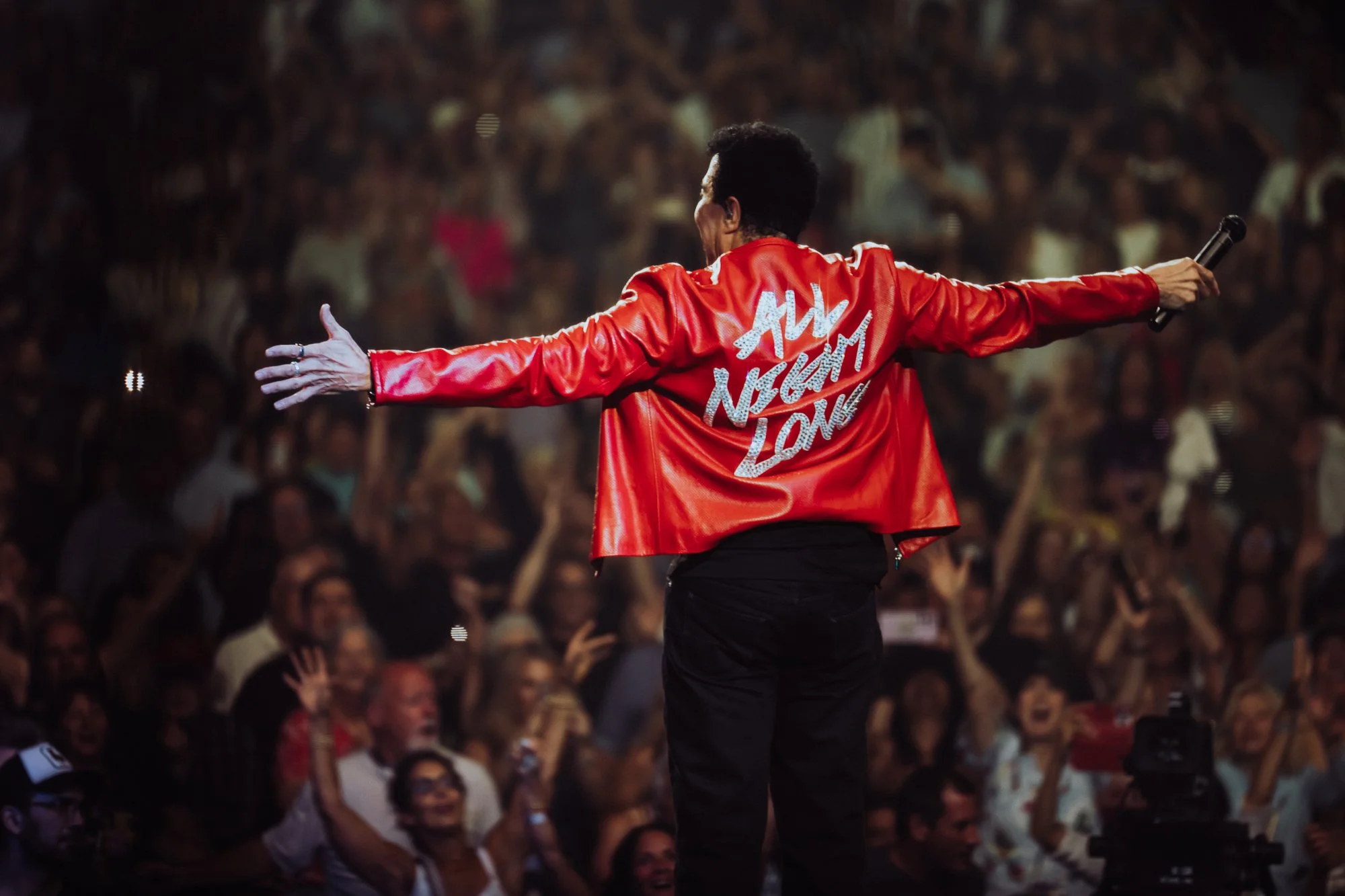 Performer on stage with arms outstretched, wearing a red jacket with white text, holding a microphone, in front of a large audience at a concert.