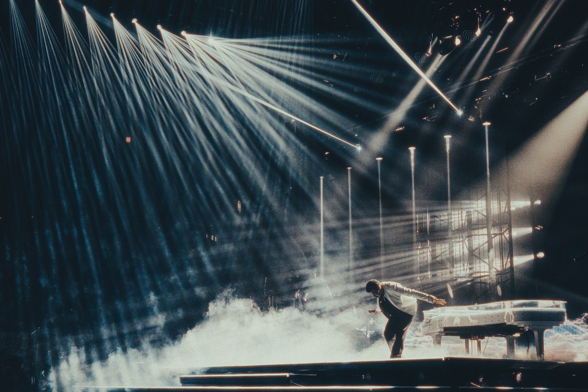 A performer lying on a table with water being sprayed over him during a live stage show under bright stage lights and fog.
