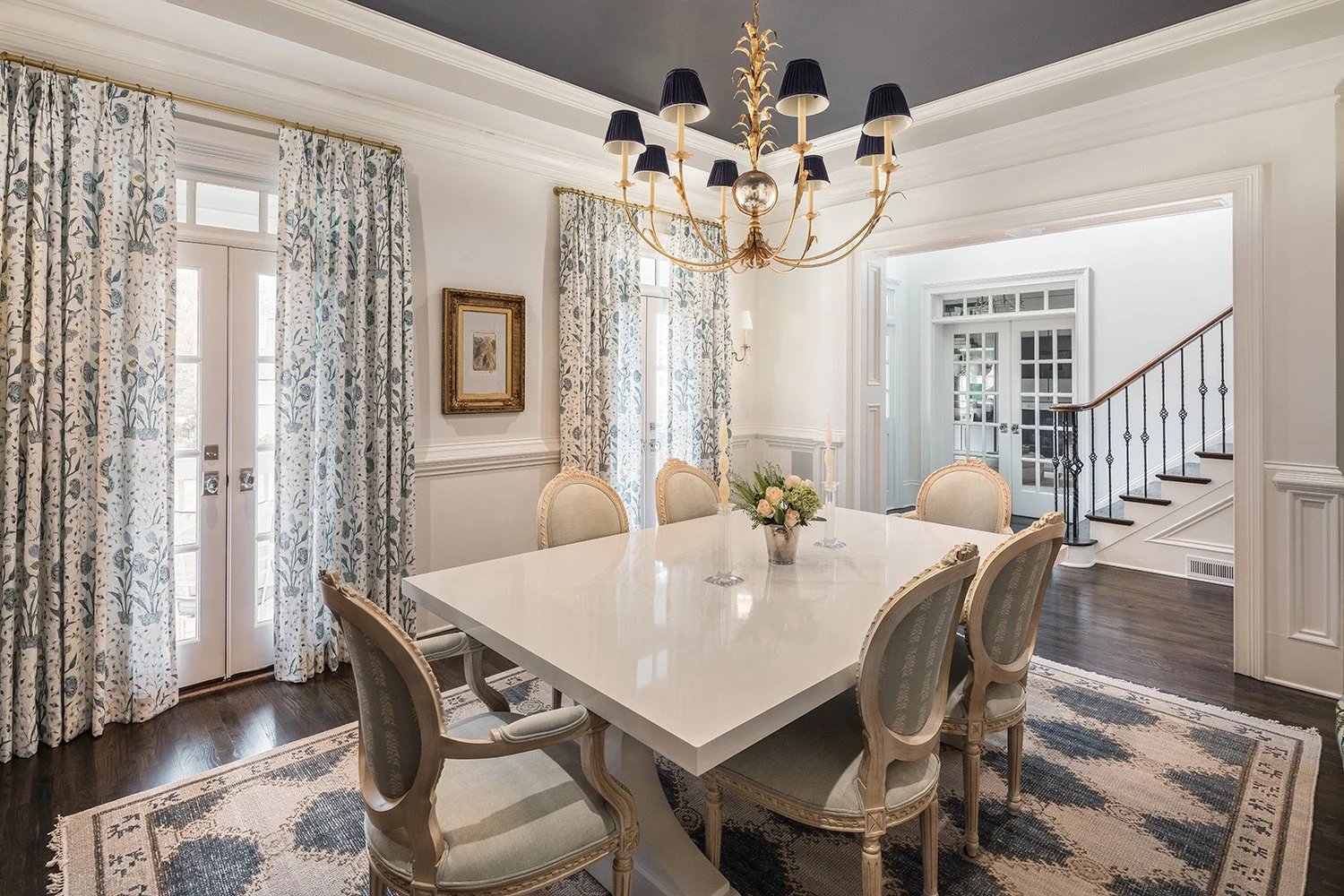 Elegant dining room with a white table, six upholstered chairs, floral curtains, a chandelier with multiple black lampshades, a floral centerpiece, and staircase with black railing.