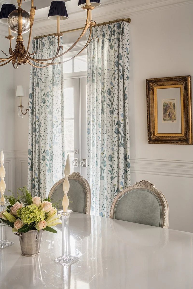 Elegant dining room with floral curtains, a chandelier, framed artwork on the wall, a white table with a floral centerpiece, and upholstered chairs.
