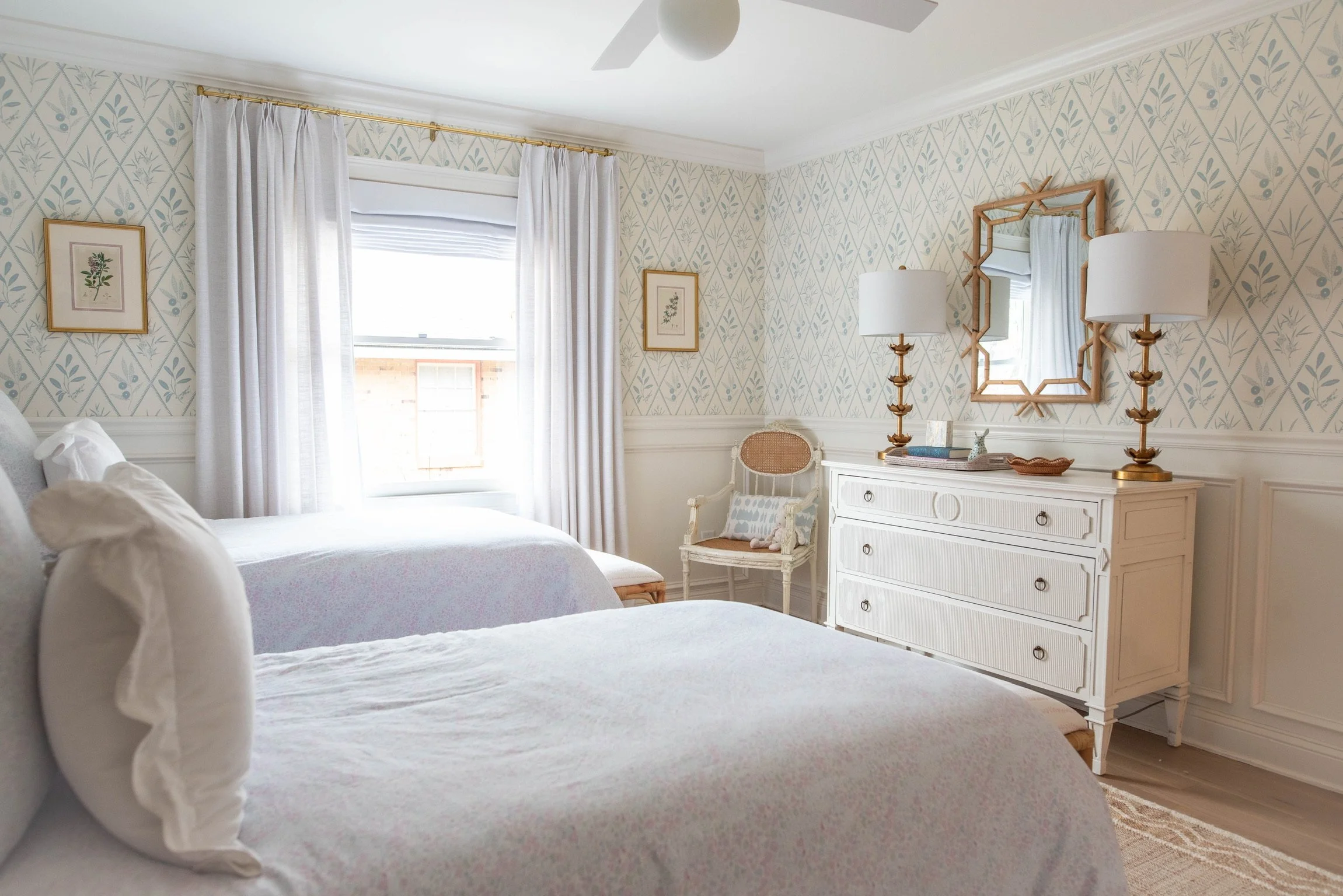 A bright, airy bedroom with two white beds, a window with white curtains, a white dresser with a mirror, and framed botanical artwork on the wall.