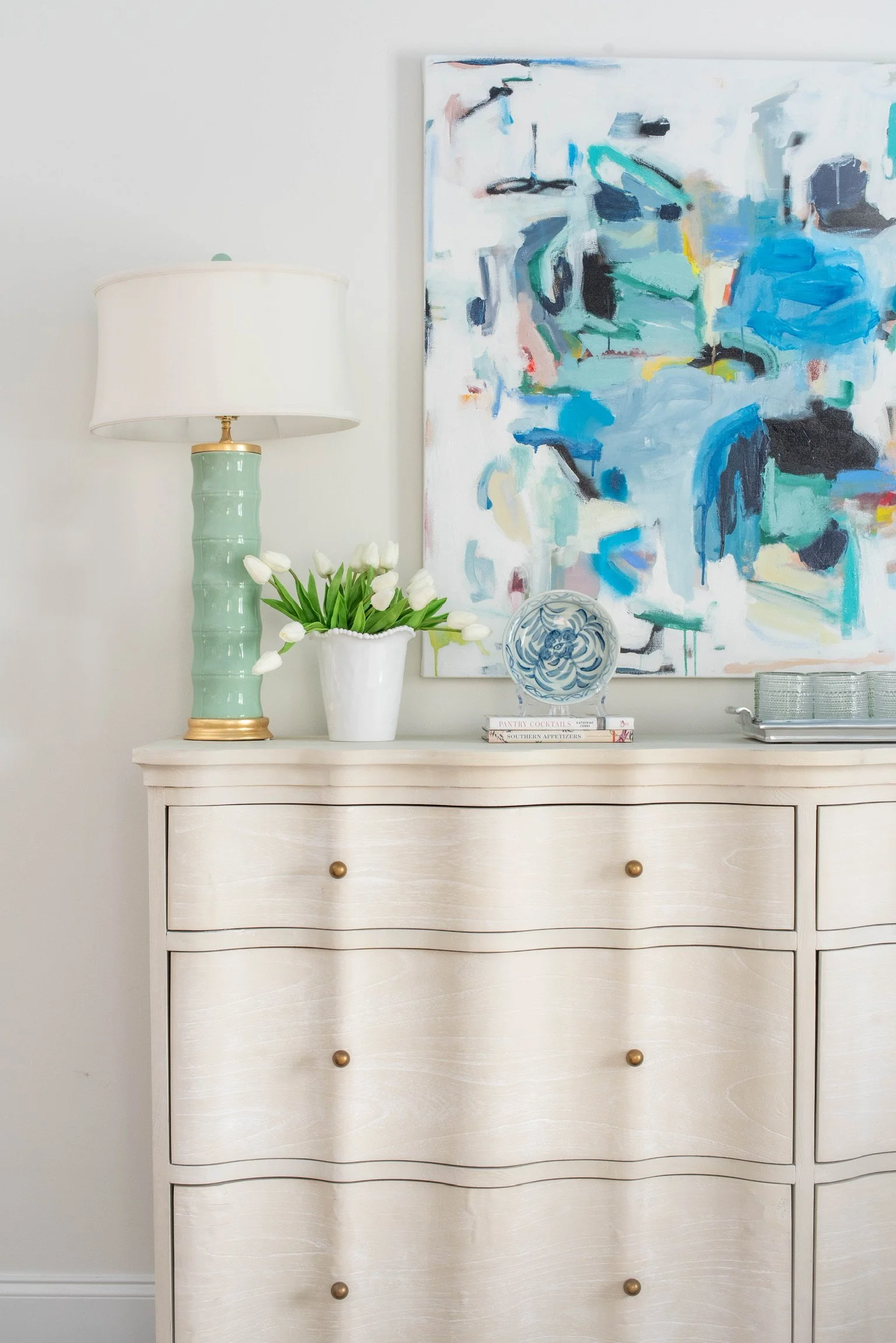 A white dresser with brass knobs decorated with a green table lamp, white tulips in a white vase, a decorative blue and white plate, and stacked books, with an abstract painting in shades of blue, green, and black hanging above.