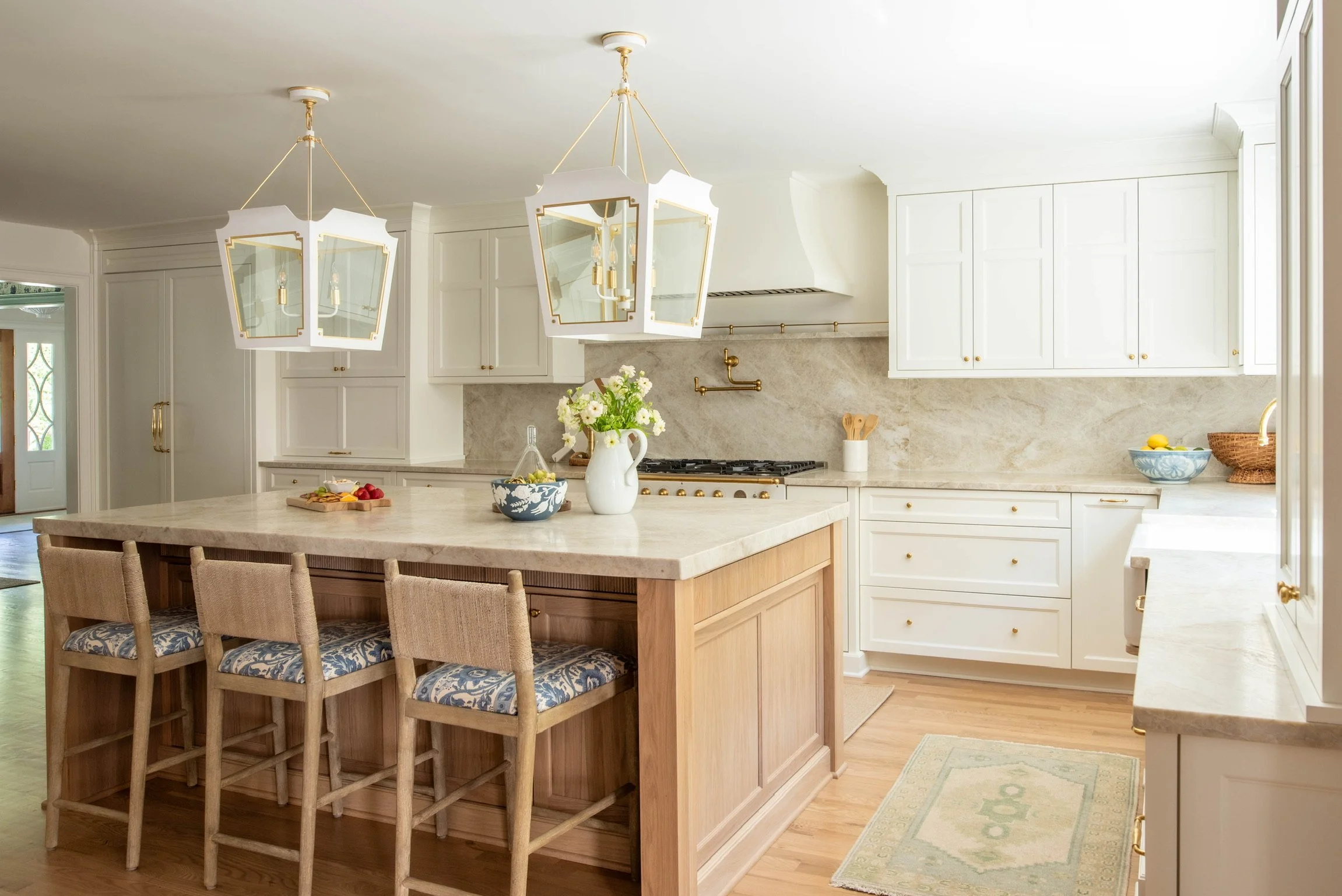 Bright kitchen with white cabinets, a large central island with a light wood base and a marble countertop, four wooden chairs with upholstered seats, three geometric pendant lights, and a window with a sink nearby; decorated with a vase of flowers, b