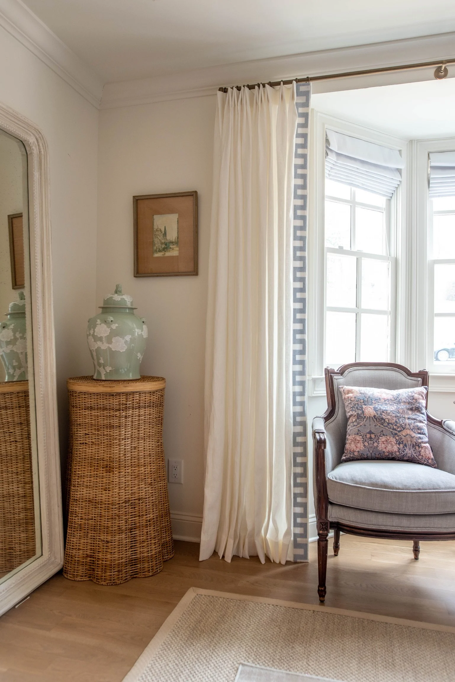 Living room corner with rattan table, jade vase, framed picture, cream curtains, and upholstered chair with floral pillow by bay window.
