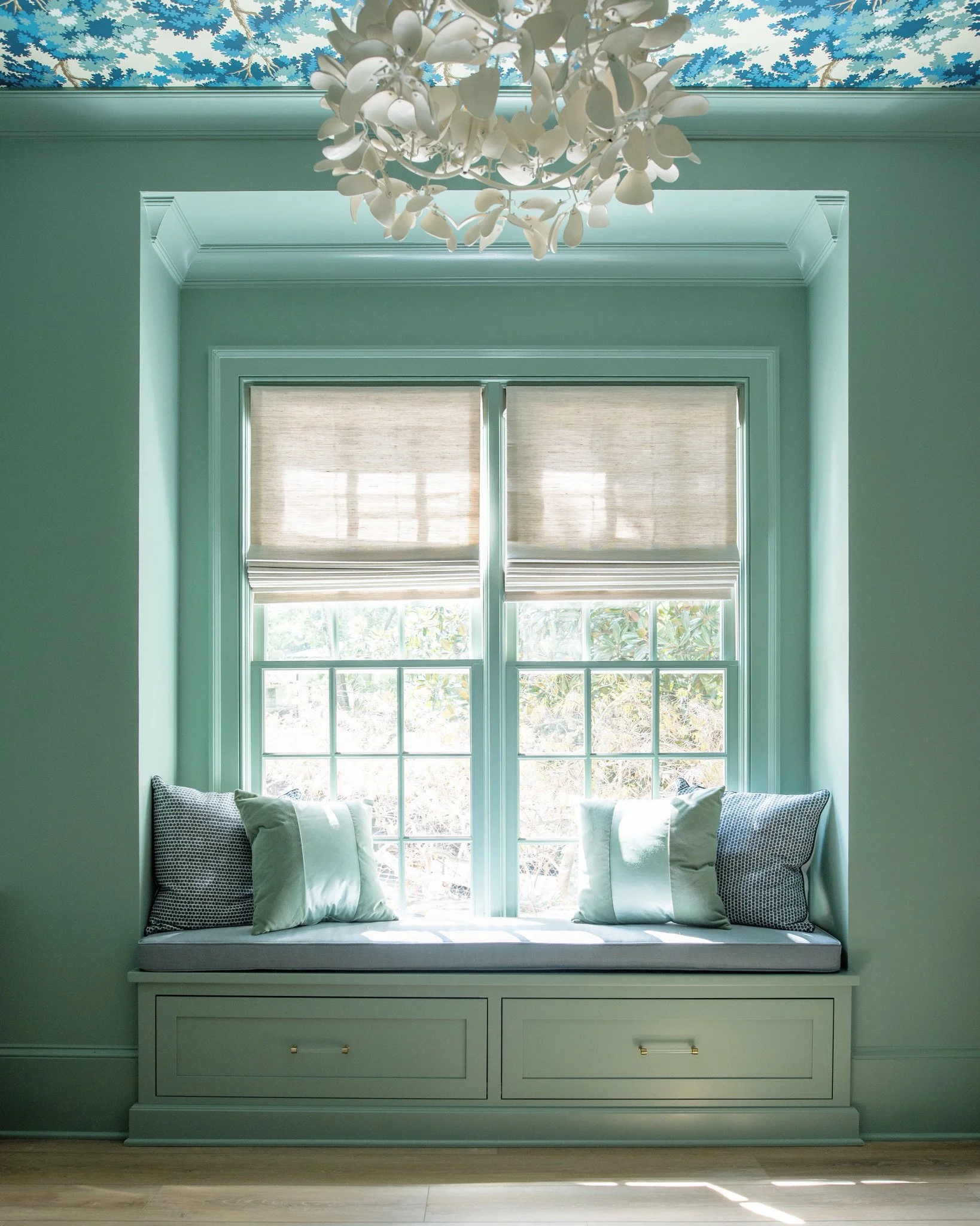 A cozy window seat with cushions and pillows in a bright, pastel-colored room, featuring large double-hung windows with beige blinds and a floral ceiling chandelier.