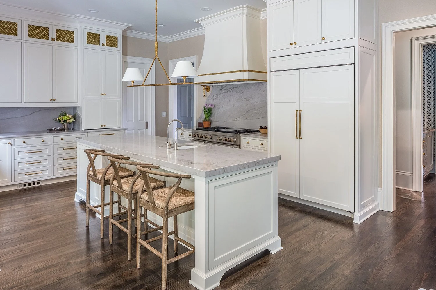 Modern kitchen with white cabinets, marble countertops, and a large island with three wooden bar stools. There is a stove with a marble backsplash, a sink, and decorative flowers on the counters. The flooring is dark wood, and the room has elegant, g