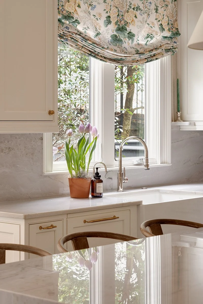 A kitchen window with floral curtains, a potted plant with pink tulips on the countertop, and a soap dispenser. The window shows trees outside, and part of a marble countertop is visible.