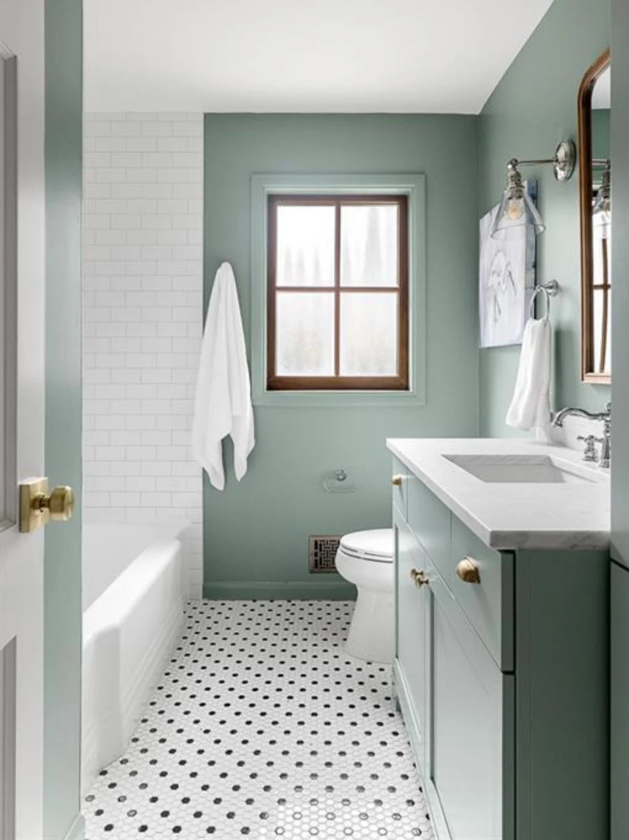 Bathroom with a window, white sink and cabinet with gold knobs, white toilet, towel hanging on wall, and a bathtub with white tiled wall behind it, featuring a small white and black dot patterned floor.
