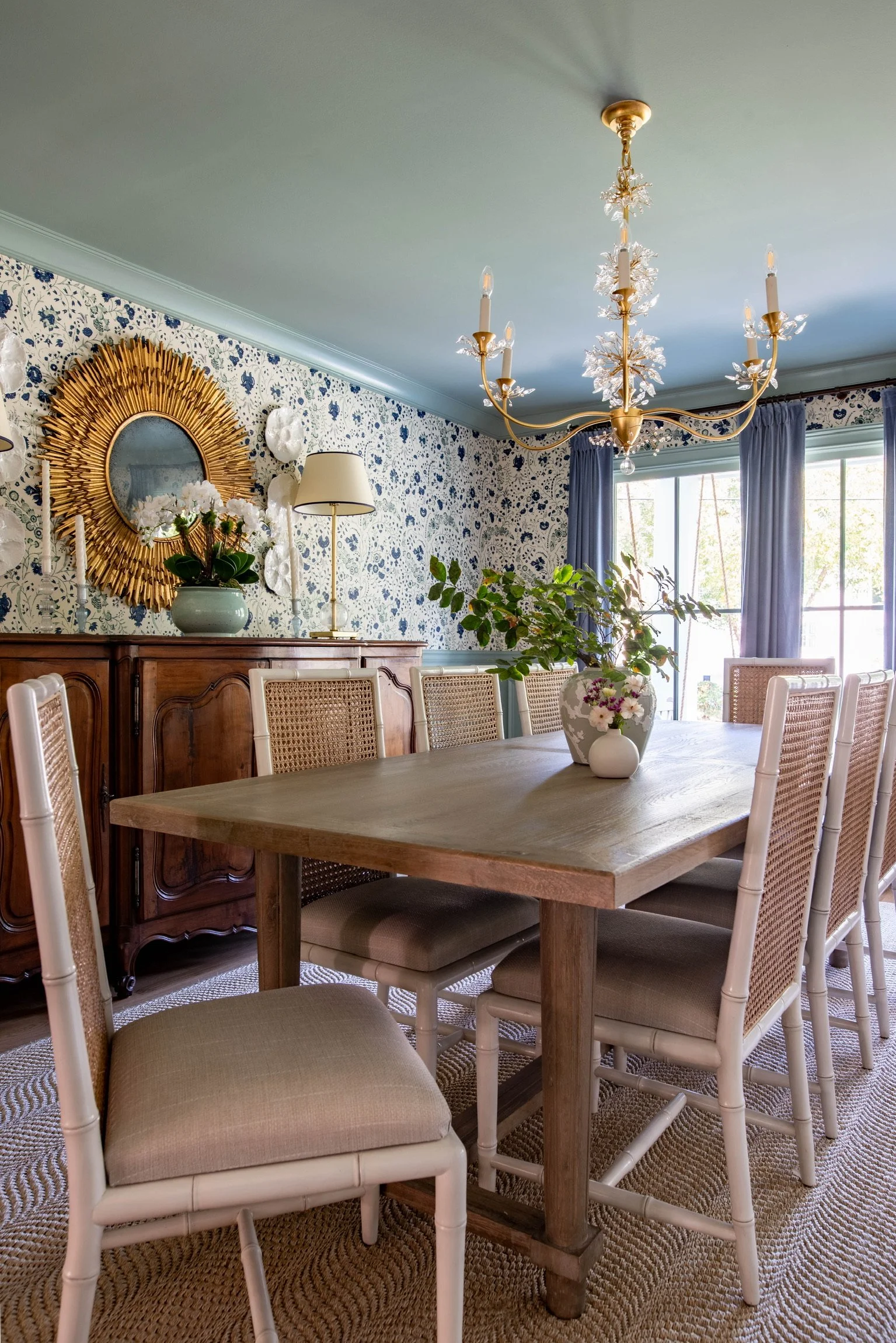 A dining room with a wooden table, eight chairs with cane backs, a wooden sideboard, a floral centerpiece, a large window with blue curtains, a gold chandelier, and decorated floral wallpaper.