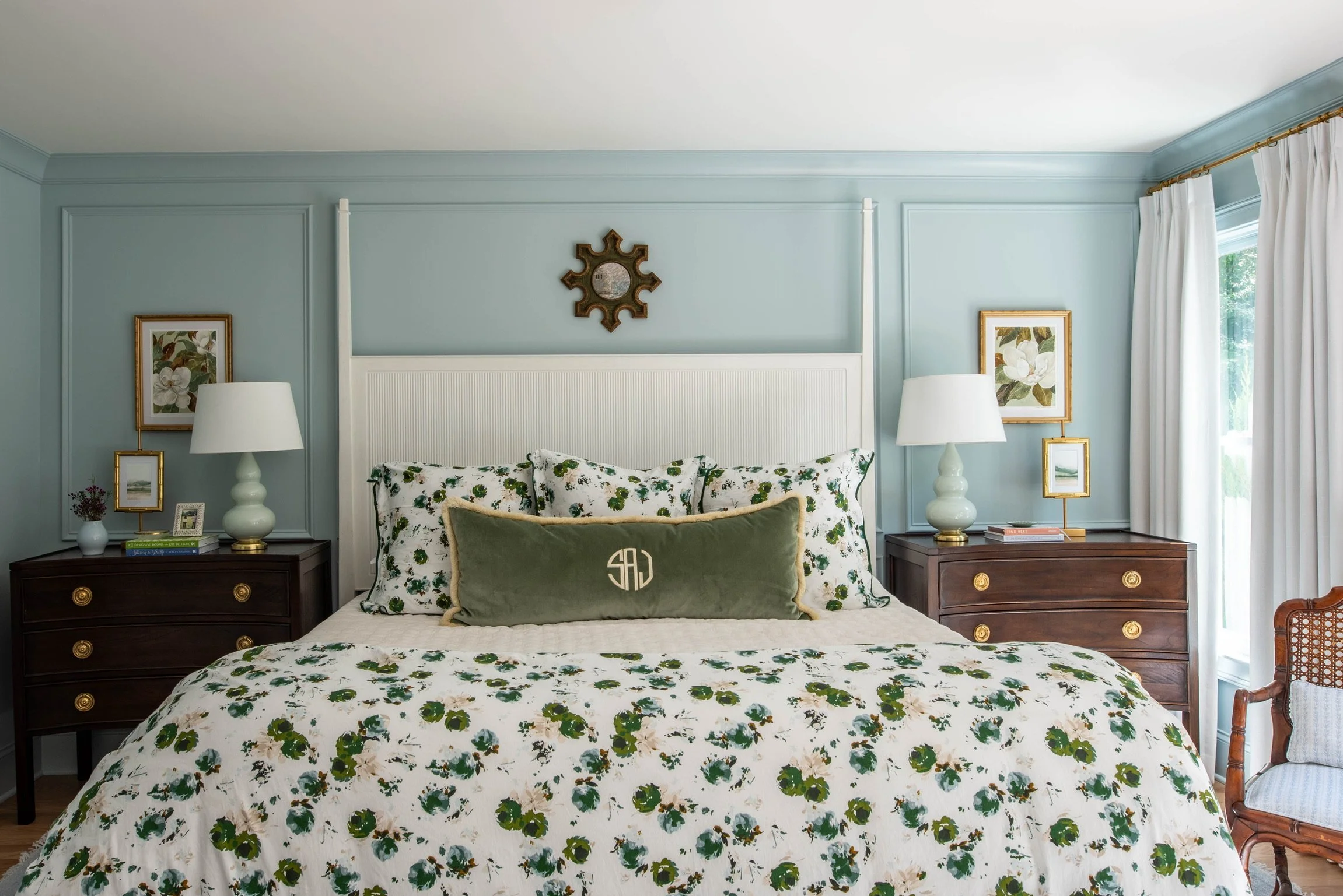 A bedroom with light blue walls, a white headboard, floral bedding, two dark wooden nightstands with white lamps, framed artwork, and a large window with white curtains.