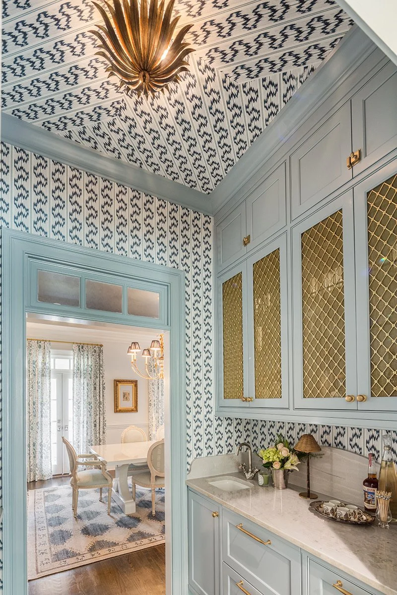 View of a kitchen with blue cabinetry, marble countertop, and patterned wallpaper; adjacent dining room with large window, curtains, and chandelier.