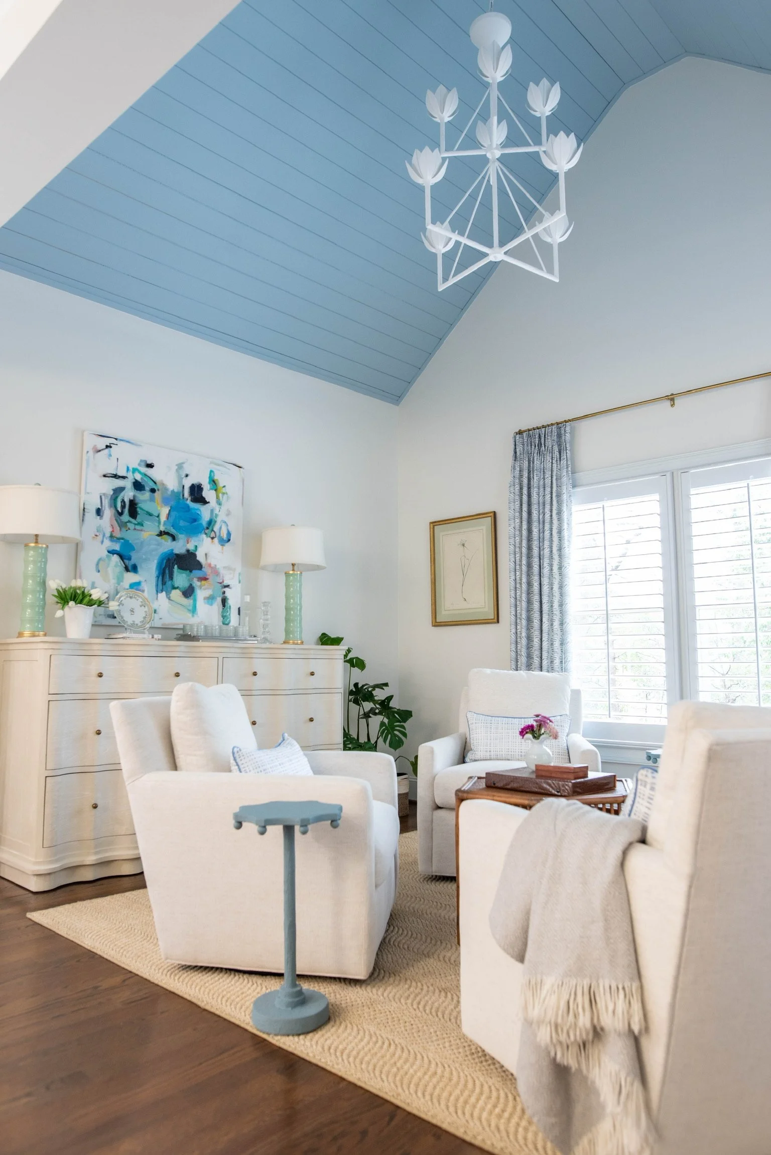 Bright living room with white armchairs, a beige wooden sideboard, abstract artwork, a blue ceiling with a chandelier, window with curtains, and a beige rug.