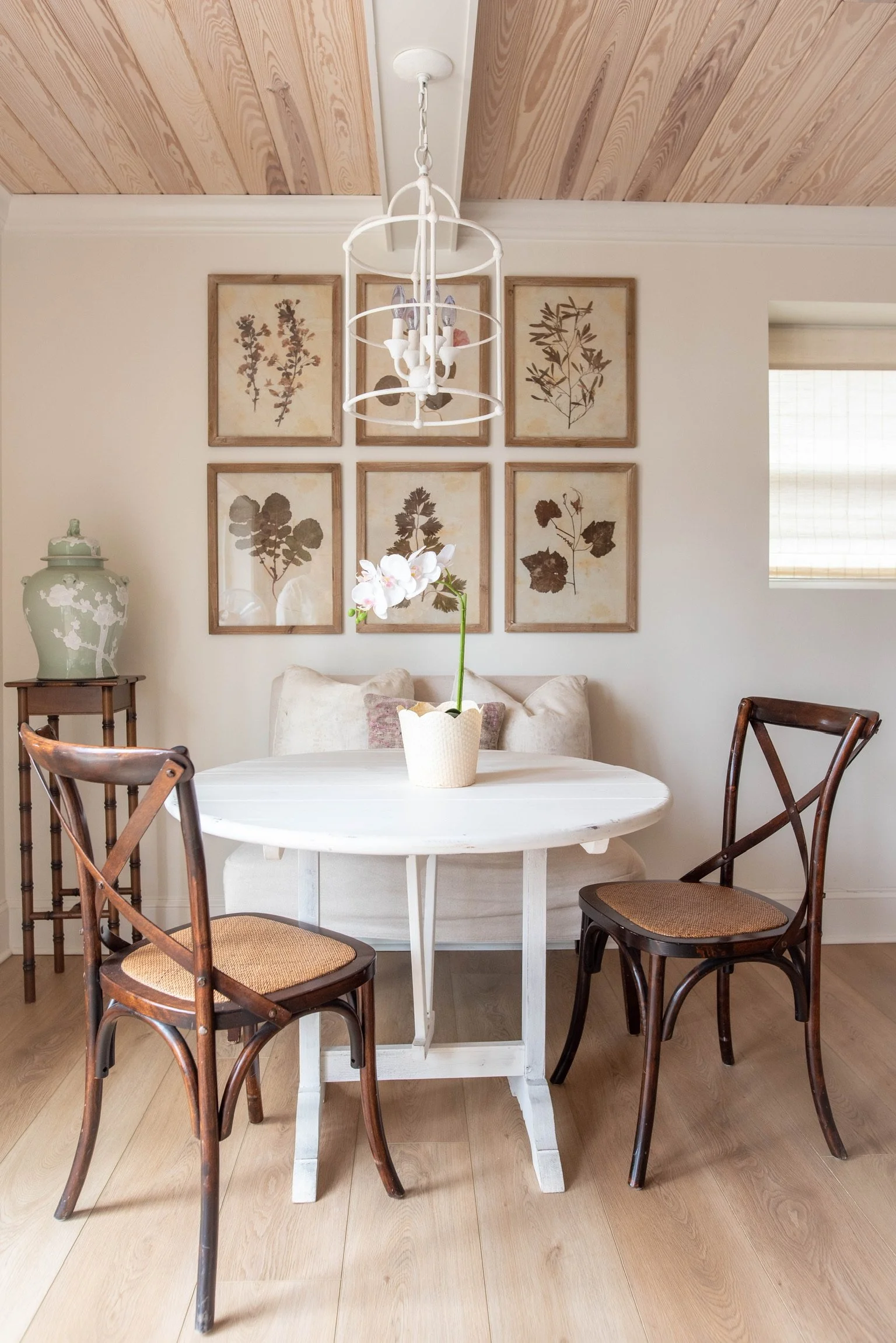 A cozy dining nook with a round white table, two wooden chairs with woven seats, a white cushioned bench with pillows, a table with a large decorative green and white vase, a white orchid in a basket, and wall art featuring pressed plant prints.