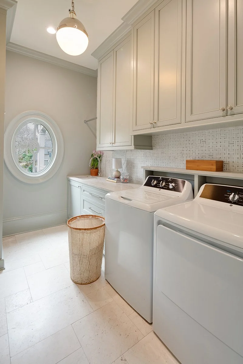 A laundry room with white cabinets, a washer and dryer, a round window, and a top of a laundry basket in the foreground.
