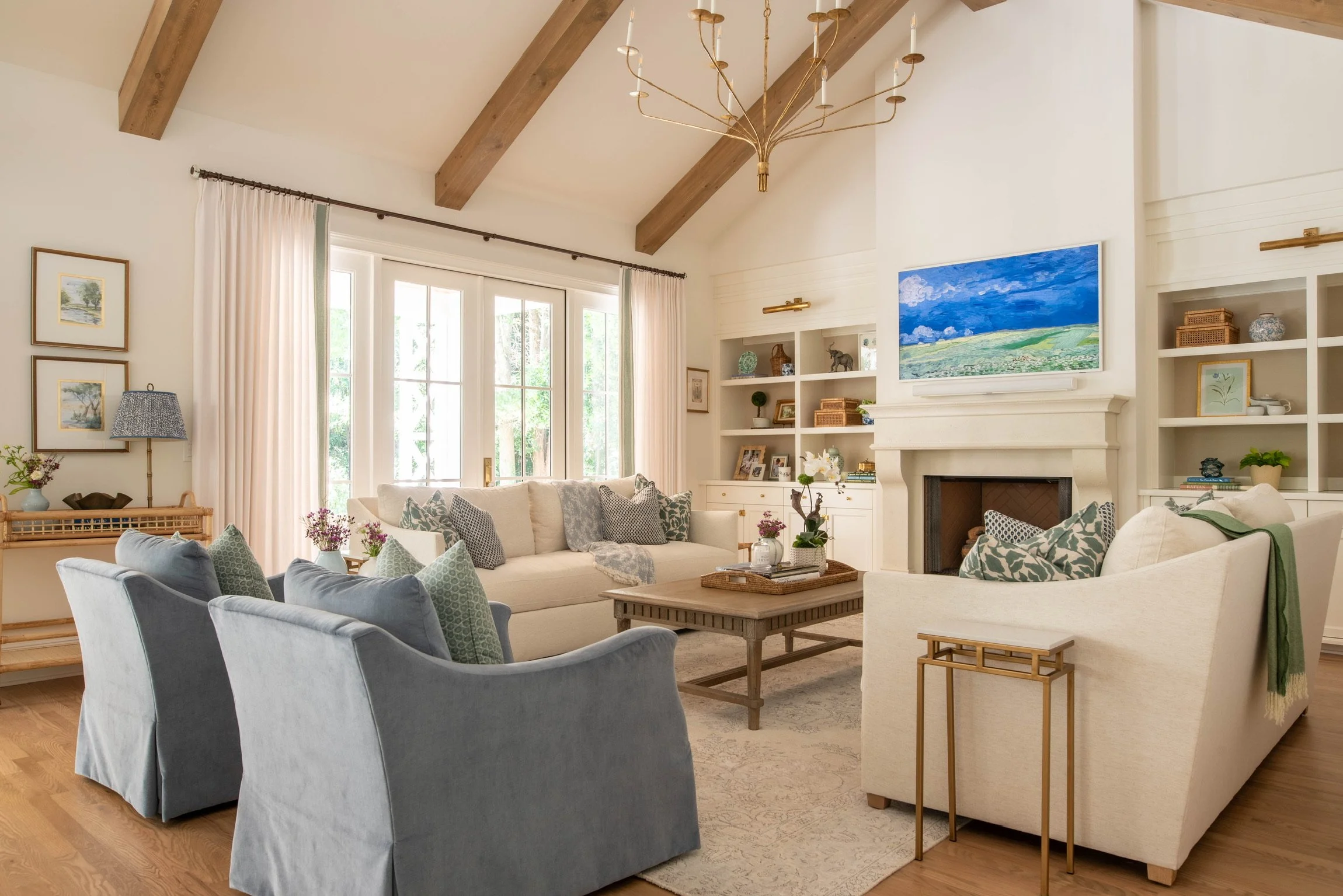 Bright living room with cream sofas, a fireplace, built-in shelves, large windows with white curtains, wooden beams on the ceiling, and a gold chandelier.