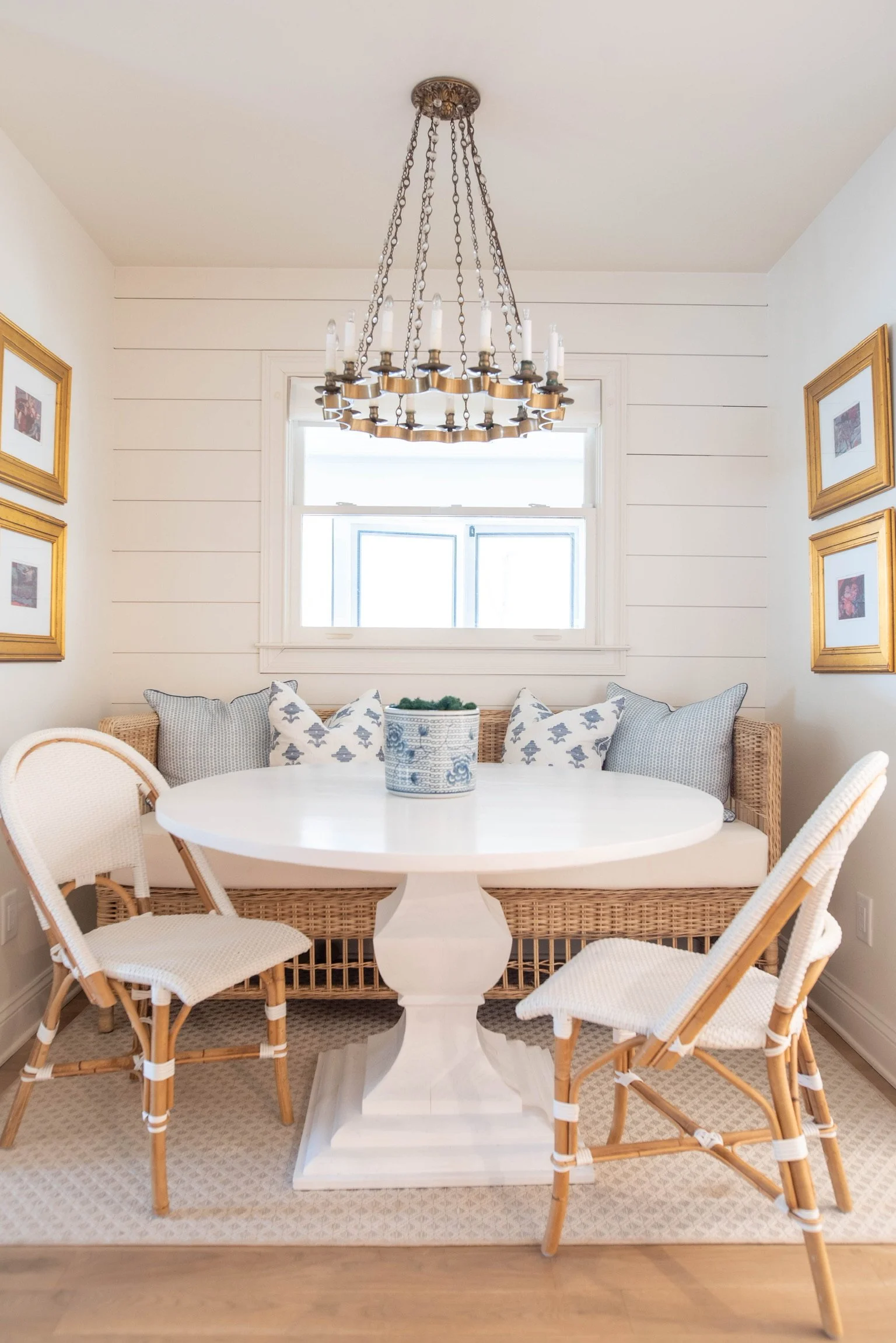 A sunlit dining nook with a white round table, surrounded by four light-colored wicker and rattan chairs, and a built-in wicker bench with patterned cushions. A window with white trim is centered above the seating, and a brass chandelier with candle-
