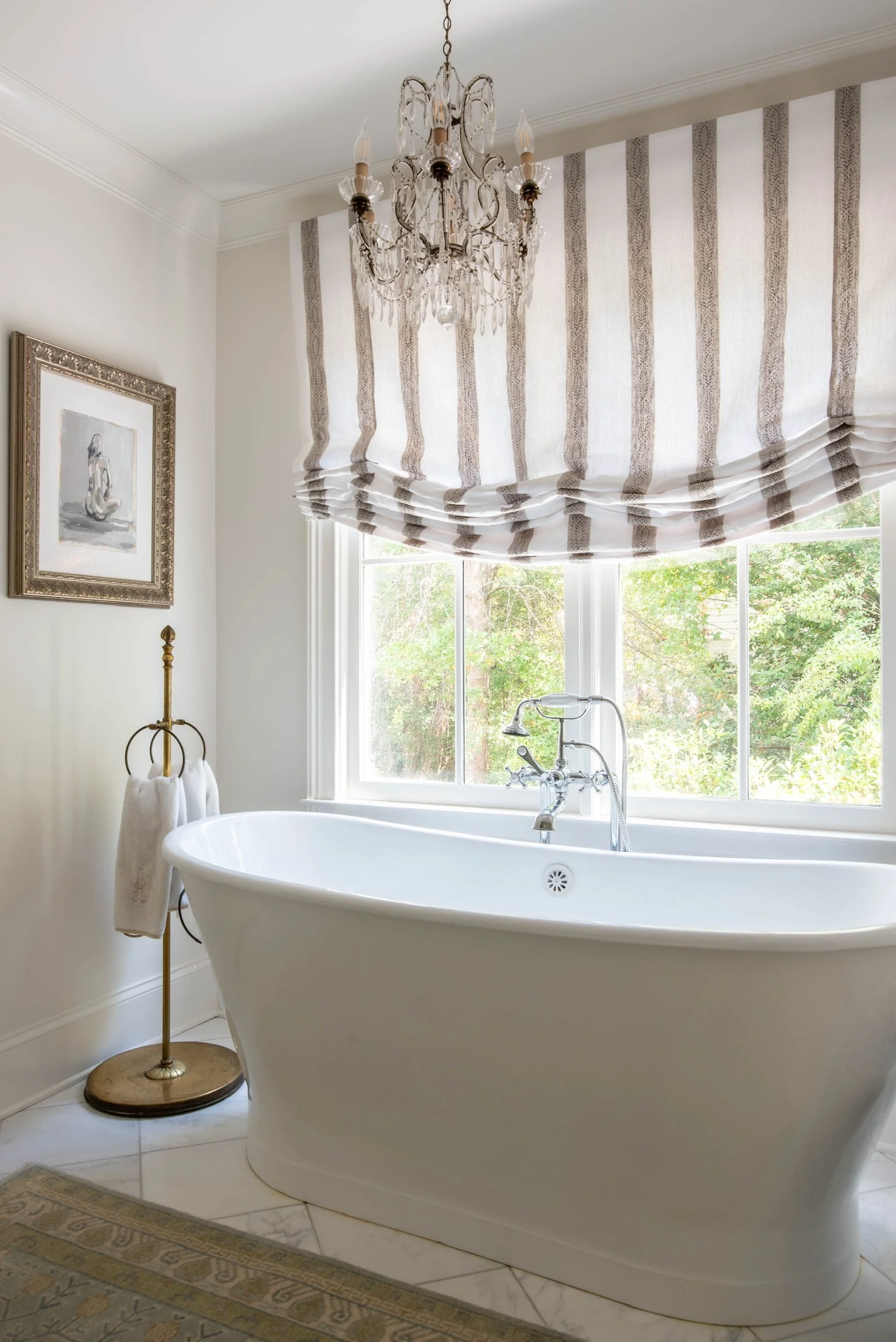 A bright bathroom with a freestanding bathtub in front of a large window with a striped Roman shade. A chandelier hangs from the ceiling, and a framed artwork is on the wall. A towel stand with towels is beside the tub, and the floor is tiled.