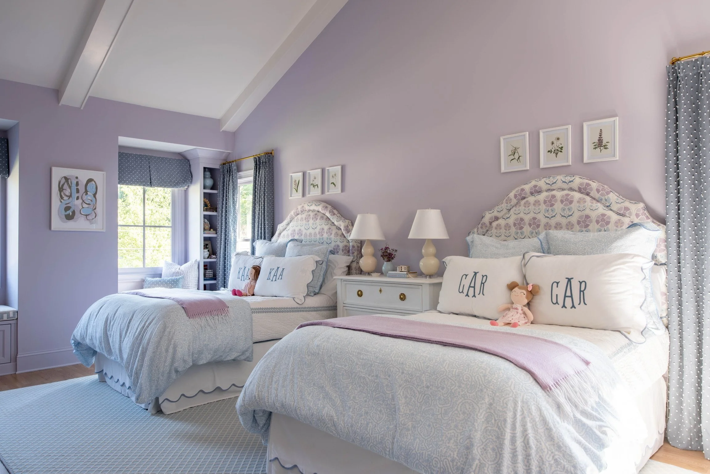 A bedroom with two beds, lavender walls, and pastel decor. Each bed has a headboard, a pillow with the words 'GAB' and 'KAA,' and small dolls. There are nightstands with lamps between the beds and framed botanical art on the wall. The room has a wind