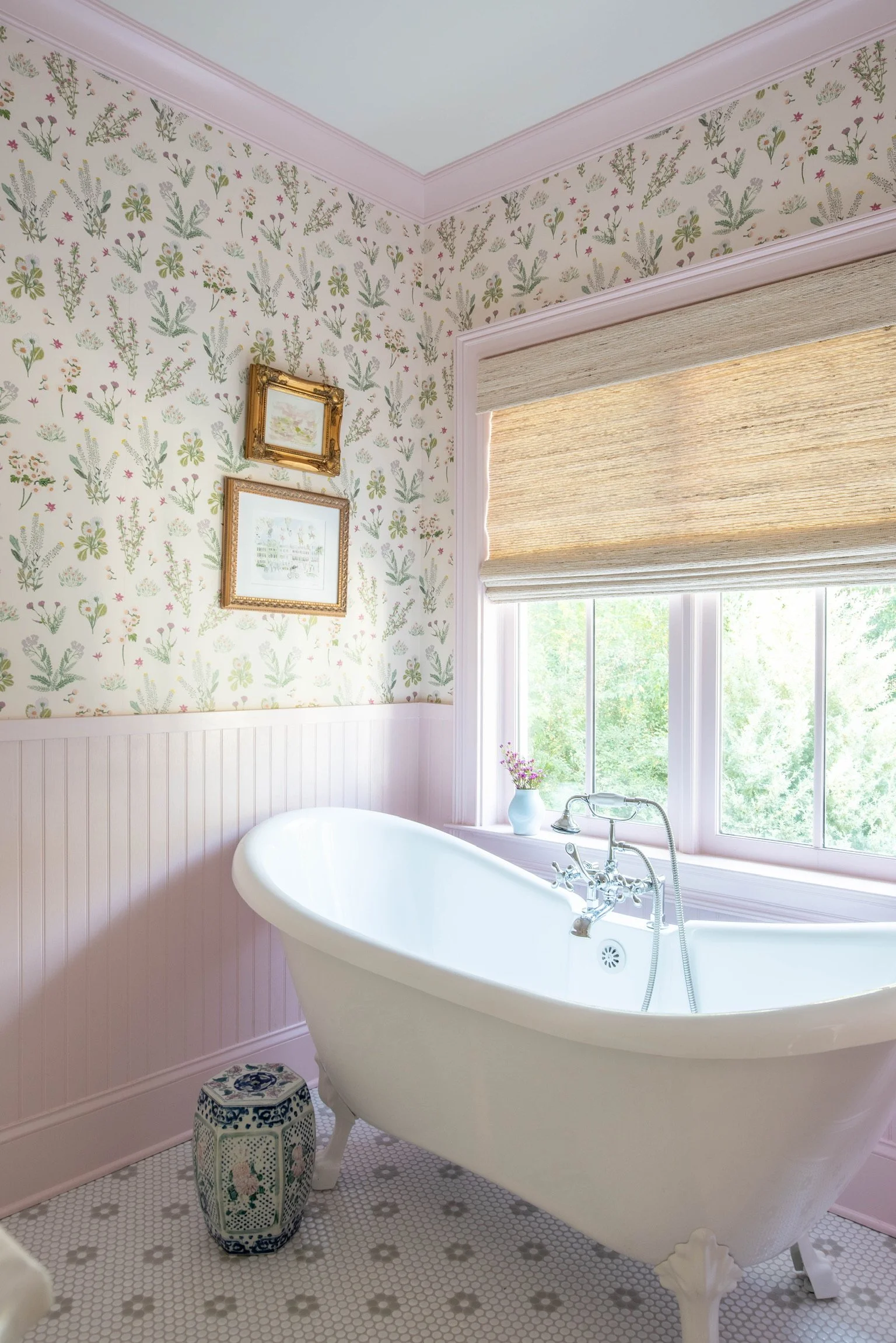 Bright bathroom with a vintage clawfoot bathtub near large window with woven shades, floral wallpaper, framed artwork, and a decorative ceramic stool.
