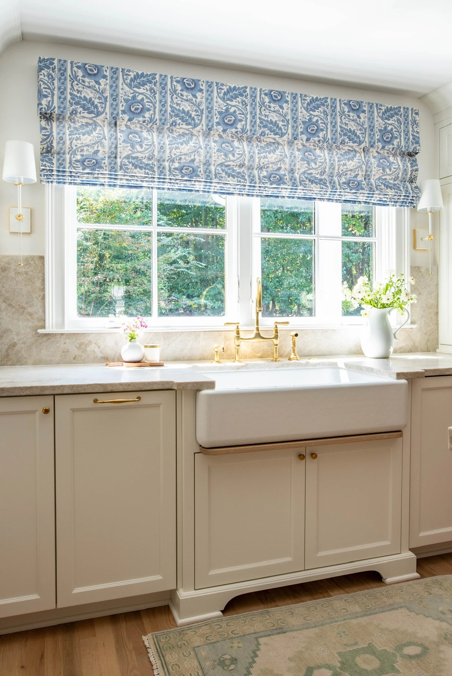 Kitchen with large window, white cabinetry, gold faucet, blue patterned window valance, and potted flowers.