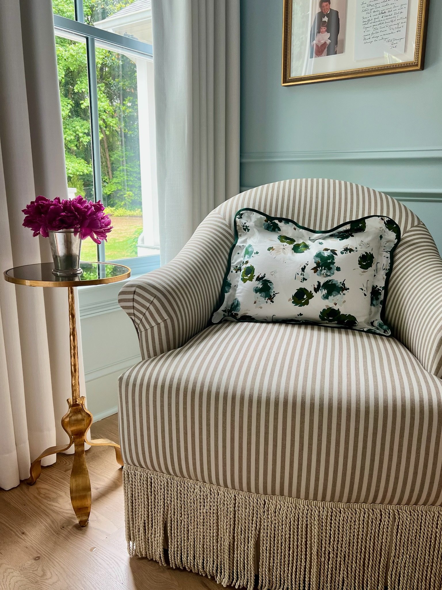 A cozy corner with a striped armchair, a floral pillow, a side table with pink flowers, and framed photographs on the wall near a window with white curtains.
