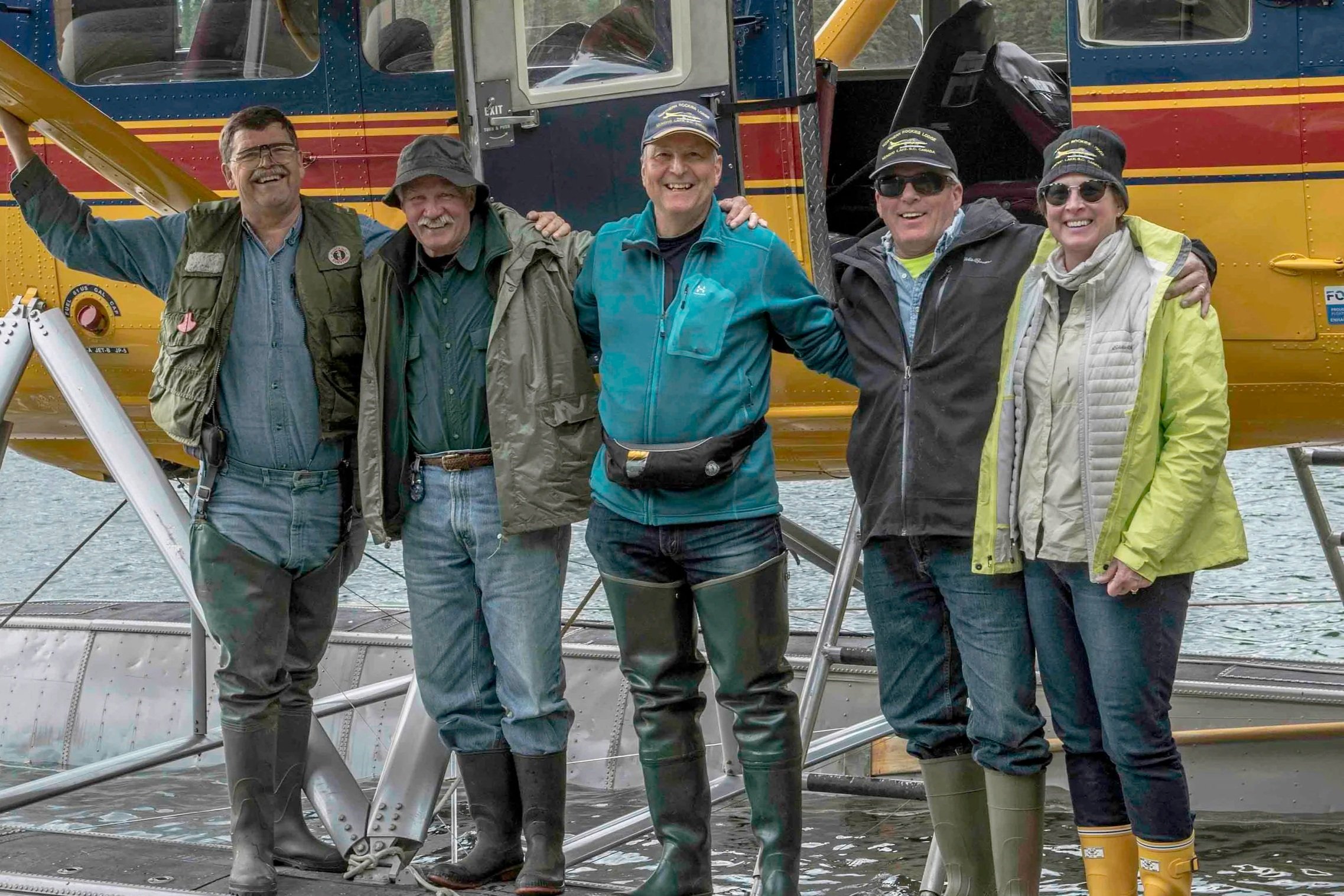 Six people standing on a dock in front of a yellow helicopter, dressed in outdoor gear and rubber boots, smiling at the camera.