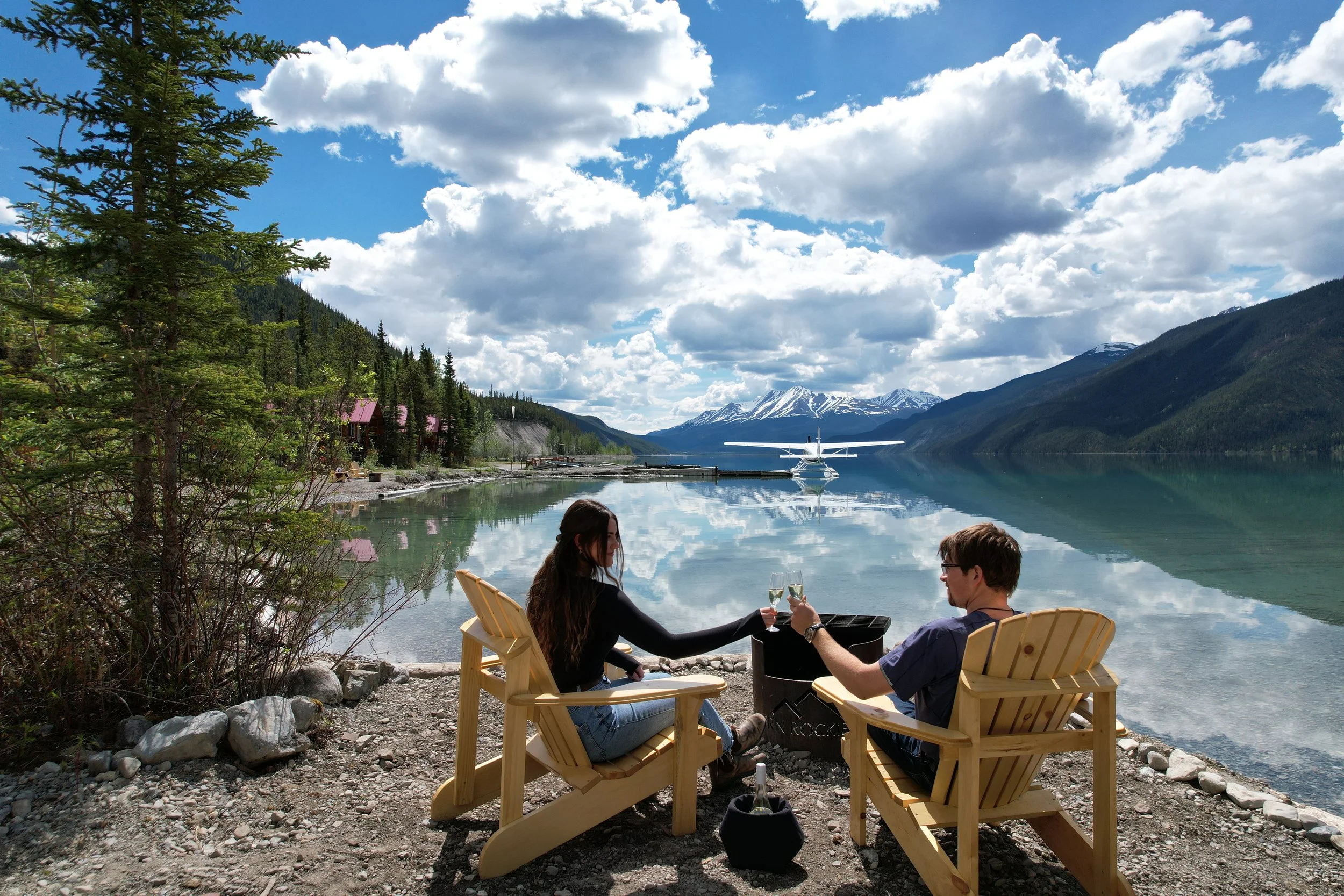 Two people sittingnat Northern Rockies lodge shoreline, drinking wine by a lake with mountains and a liard air floatplane in the background.