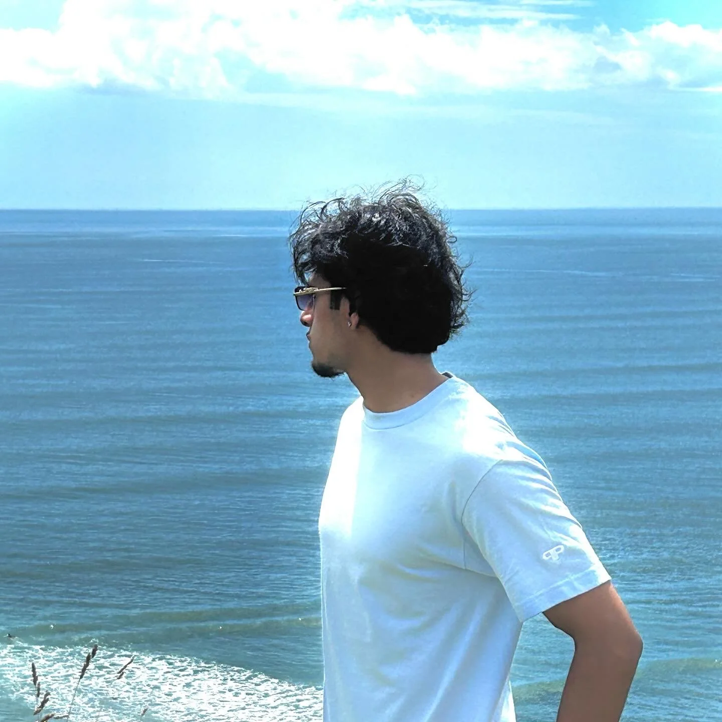 A man with curly black hair and sunglasses facing sideways stands near the ocean, with blue water and a partly cloudy sky in the background.