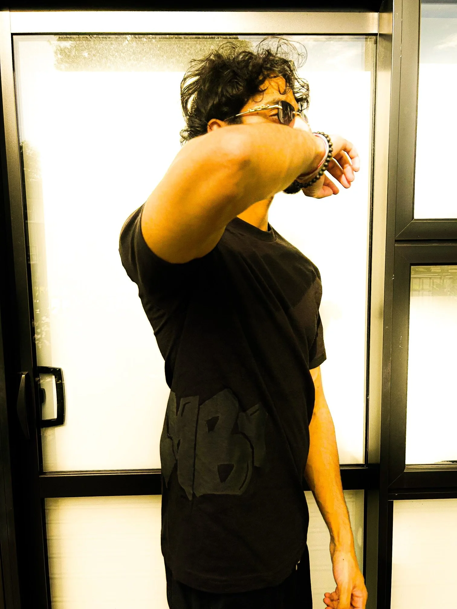 A person with curly dark hair, wearing sunglasses, a black T-shirt, and a bracelet, standing in front of an elevator, covering their face with their arm.