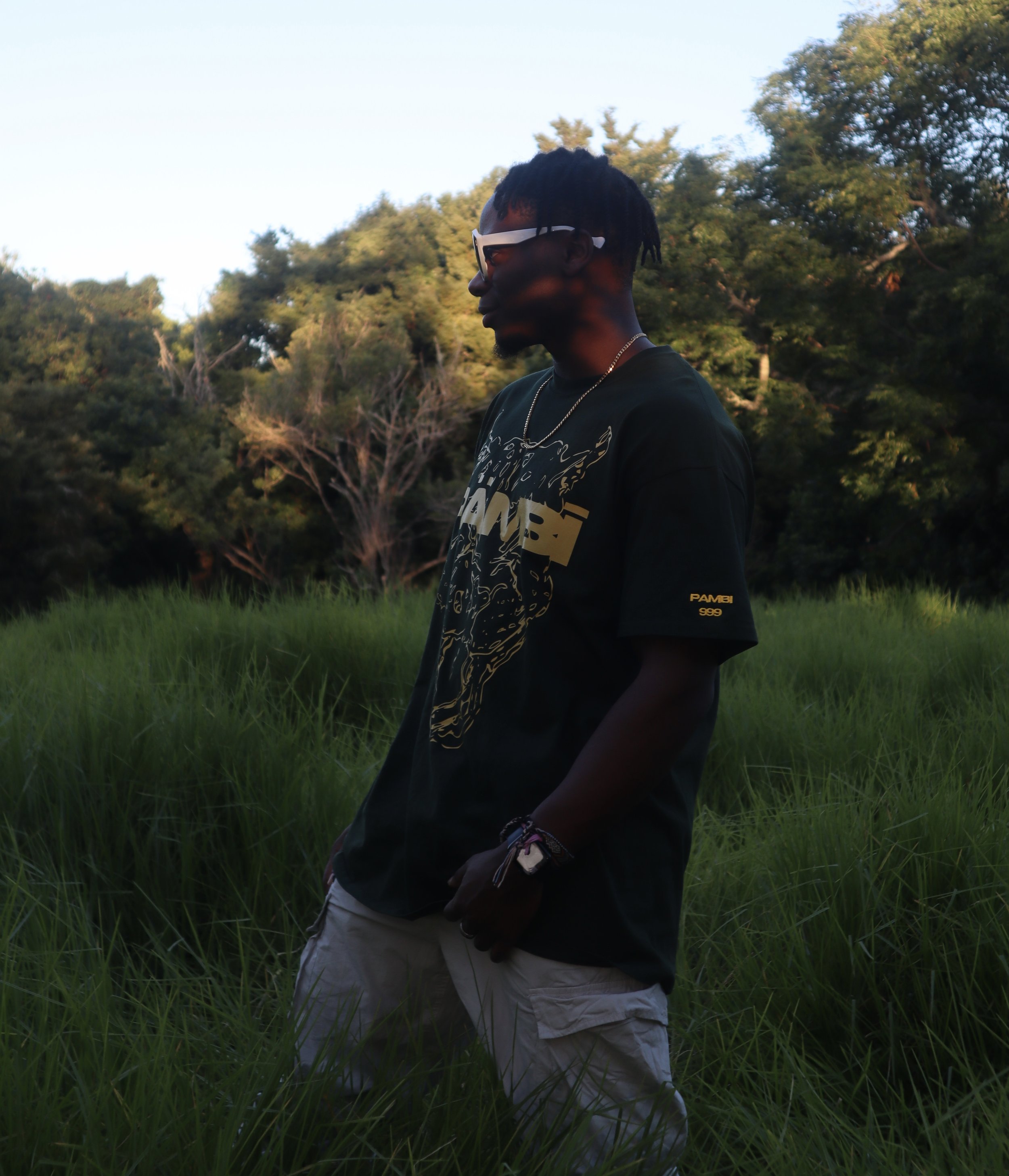 A young man stands in a grassy field with trees in the background, wearing sunglasses, a dark graphic t-shirt, shorts, and jewelry during the daytime.
