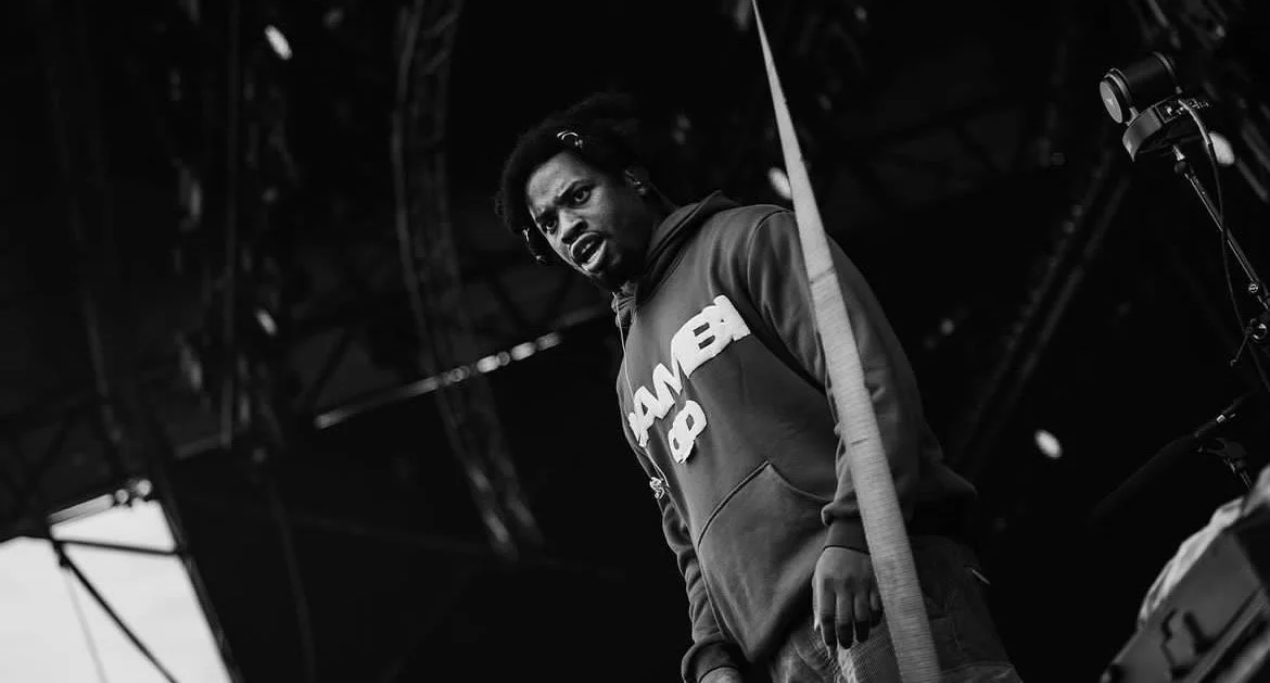 A man with dreadlocks and a beard wearing a hoodie, standing on a stage with a serious expression, under stage lighting and structure.