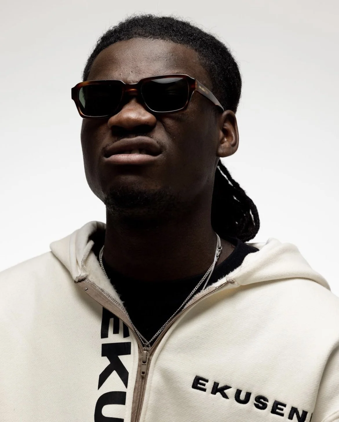 Close-up of a man with dark skin wearing black sunglasses, a white hoodie with black text, and necklaces, against a plain light background.