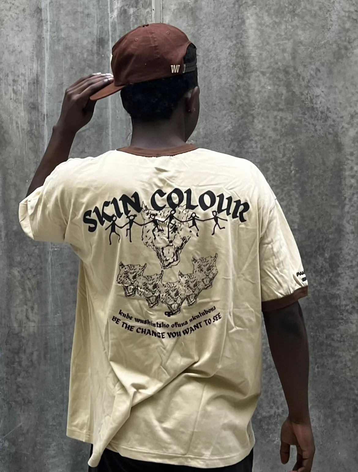A person wearing a beige T-shirt with the text 'SKIN COLOUR' and a graphic of a lion's head and three leopards, along with the phrase 'BE THE CHANGE YOU WANT TO SEE'. The person is also wearing a brown cap and is standing against a plain concrete wall, facing away from the camera.