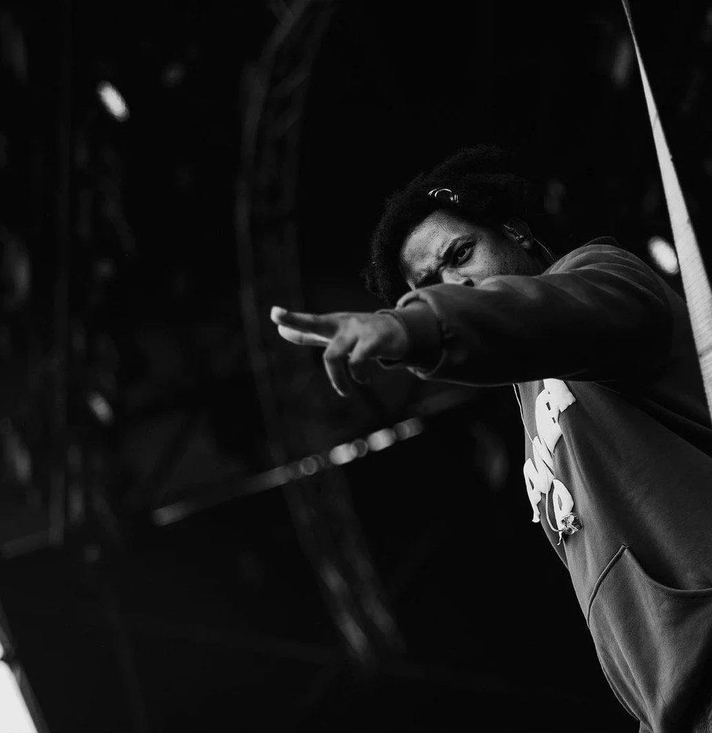 A person with dark curly hair, wearing glasses, is pointing towards the camera with an intense expression, against a dark background.