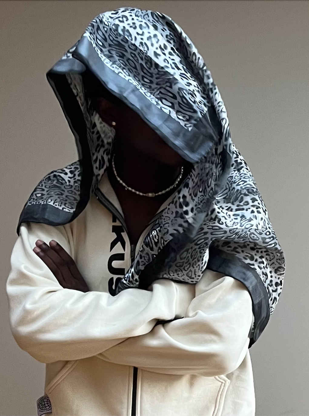 Person wearing a black and white animal print hooded jacket with matching head wrap, crossing arms, against a plain background.