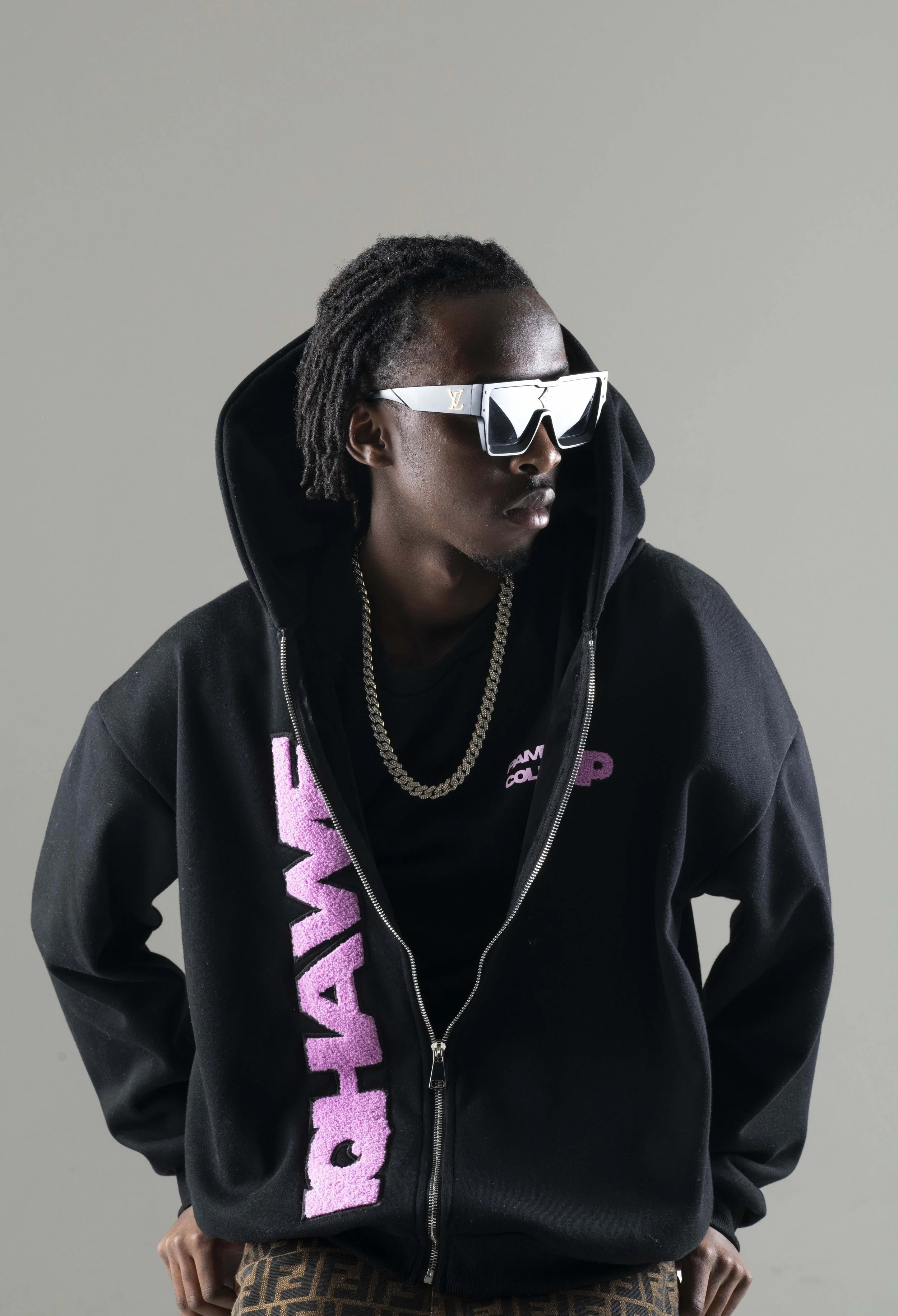 A young man with dreadlocks wearing oversized sunglasses, a black hoodie, a black T-shirt with pink text, gold chains, and patterned pants, standing against a plain background.