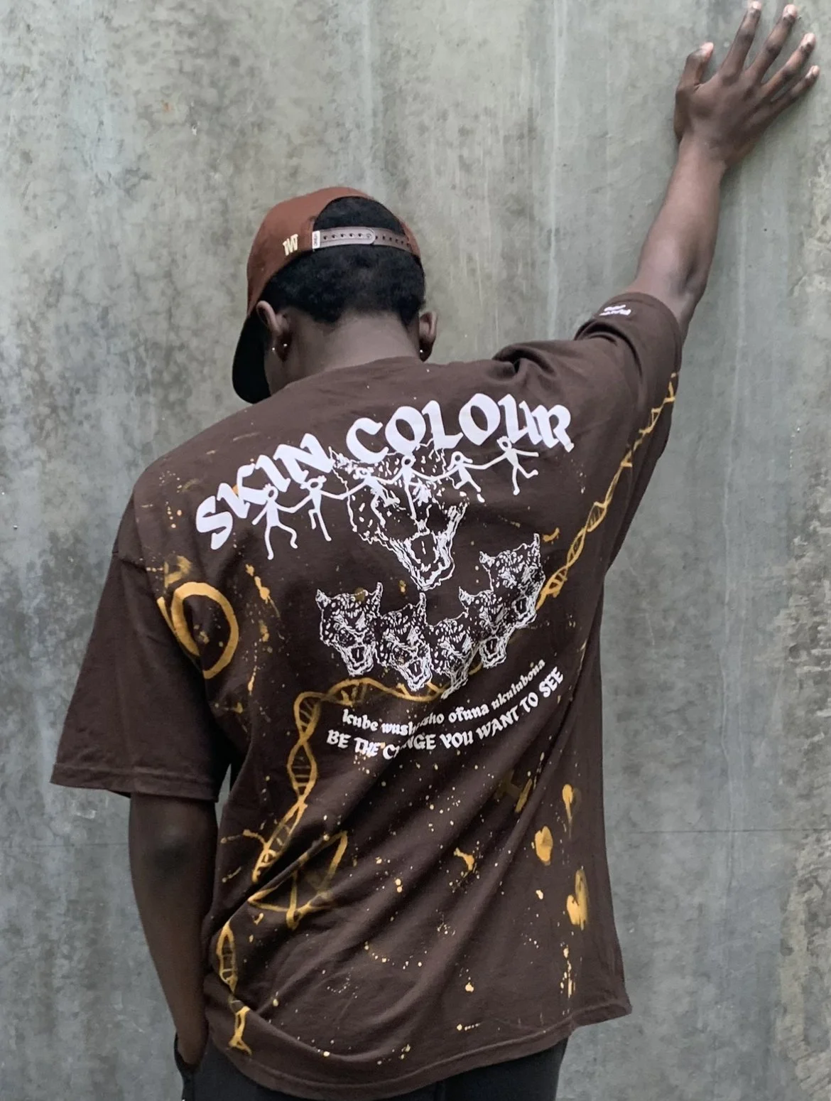 Person with dark skin wearing a brown graphic T-shirt and a backwards baseball cap, leaning against a textured wall with one arm extended upward and hand flat against the wall.