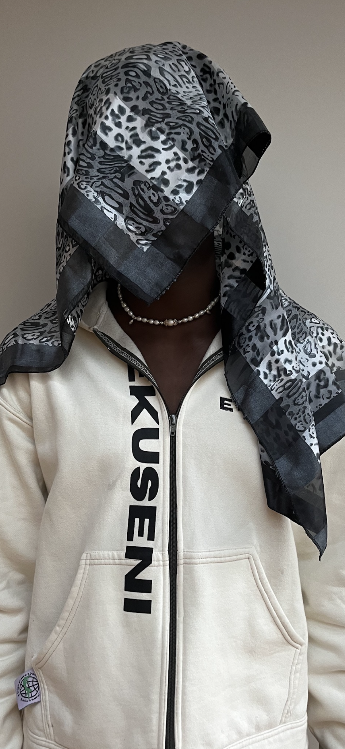 Person wearing a white zip-up hoodie with black text, a pearl necklace, and a leopard print headscarf covering their face and head.