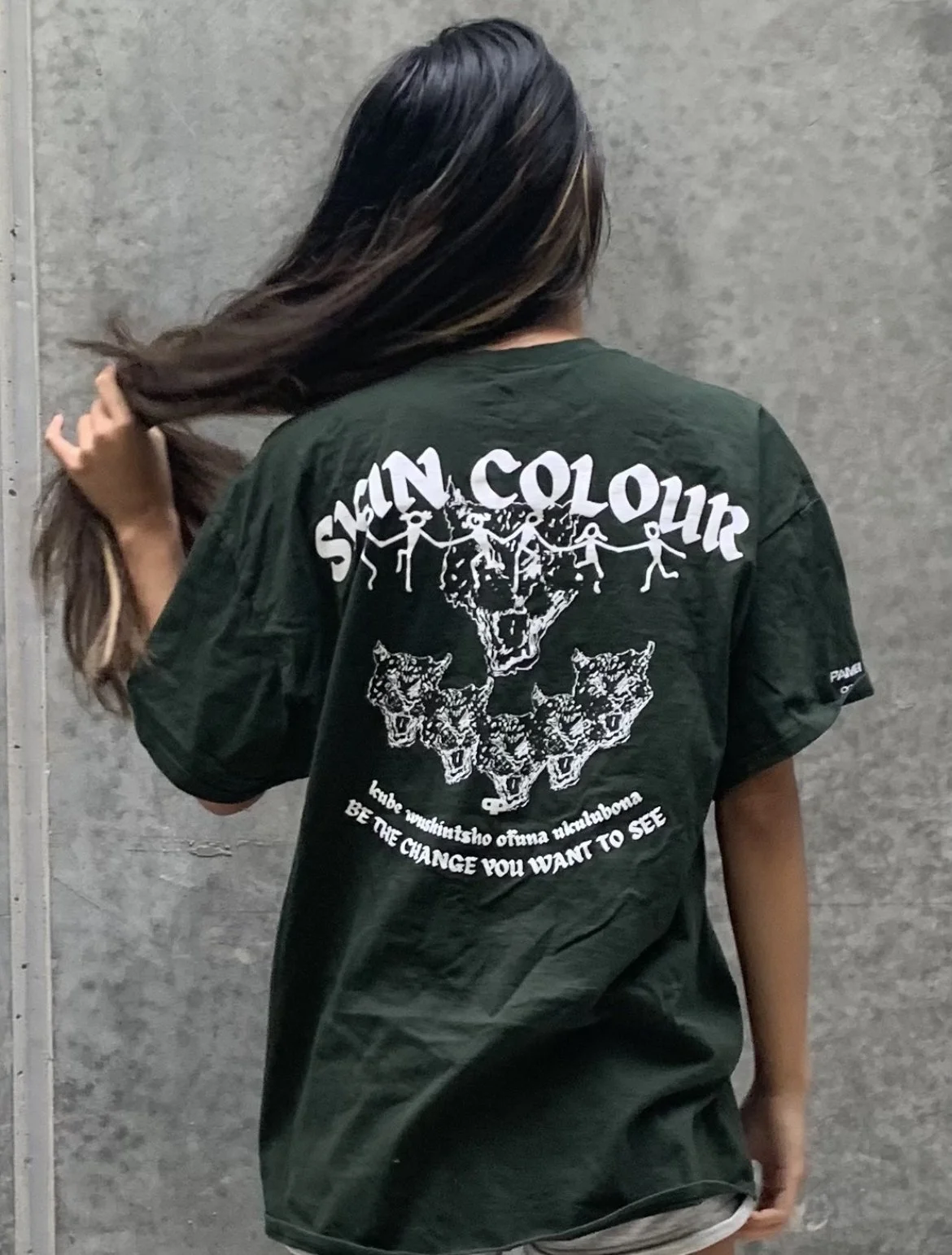 A person with long dark hair touching their hair, wearing a black T-shirt with white graphic text and images, standing against a gray industrial wall.