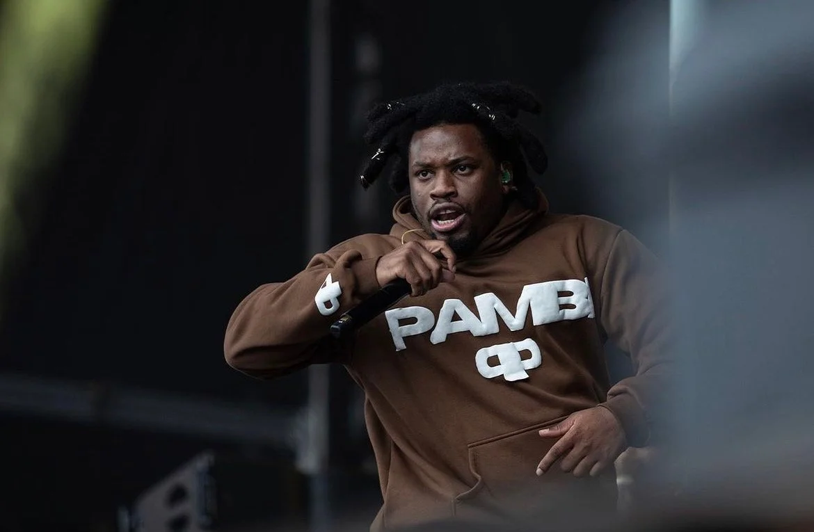A man with dreadlocks wearing a brown hoodie with the word PAME on it holding a microphone and speaking passionately.