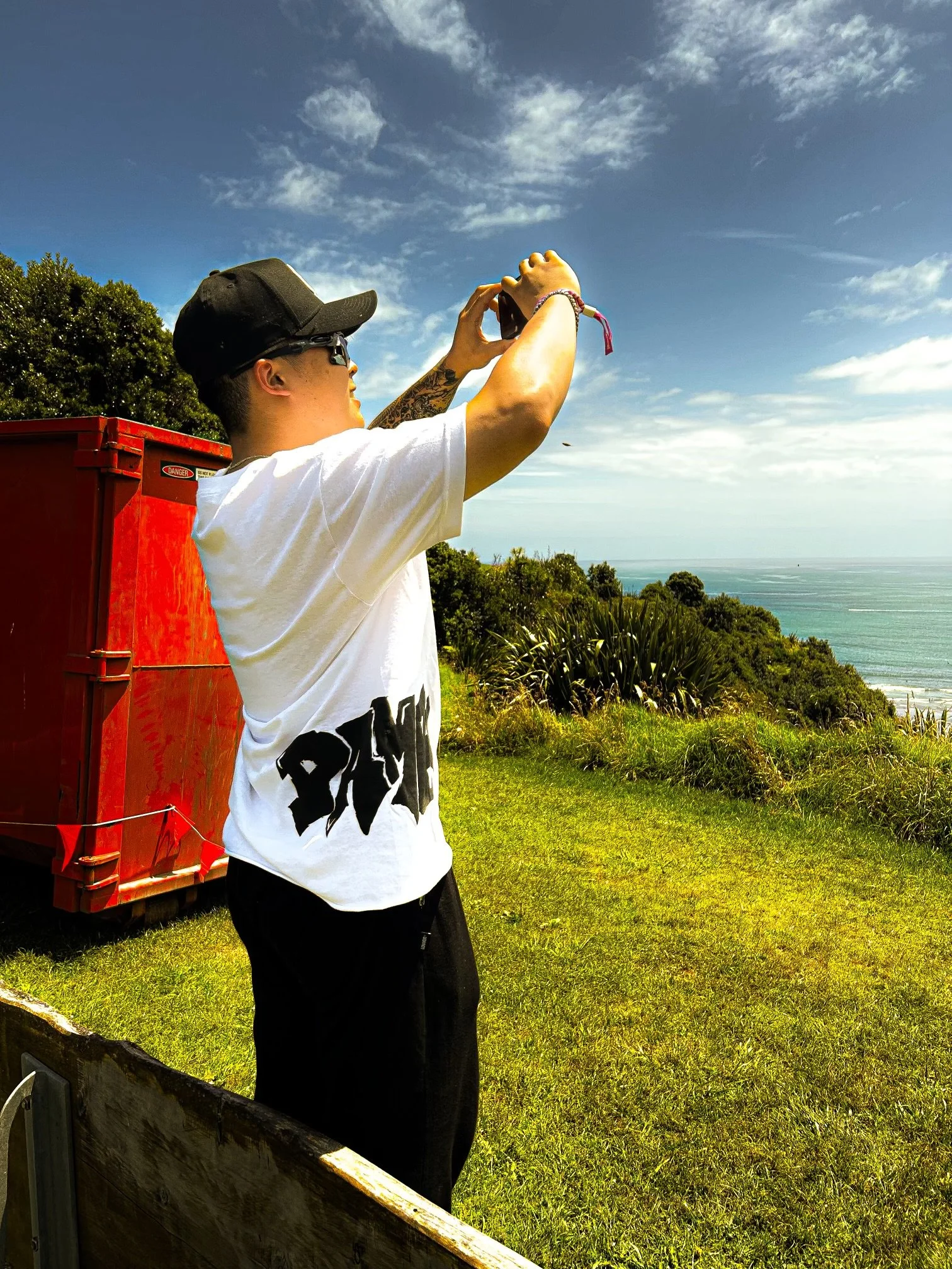 A person standing outdoors on a grassy area, wearing a black cap and sunglasses, taking a photo with a smartphone. They are near a red box, with the ocean and blue sky with white clouds in the background.