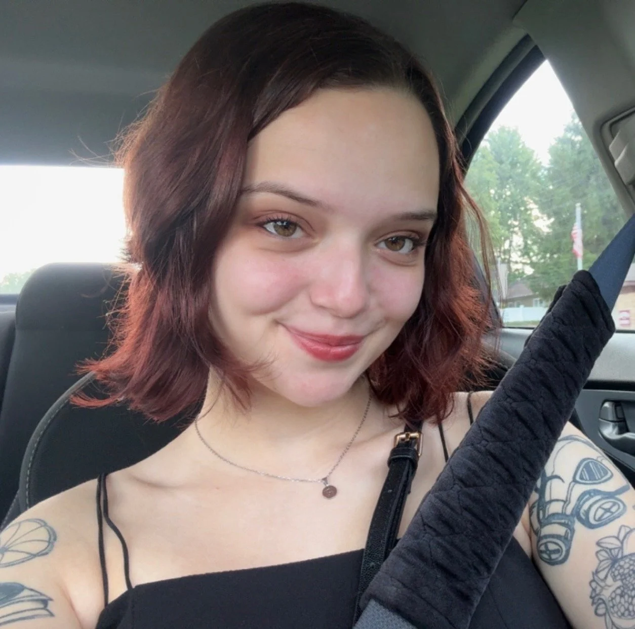 A young woman with short, wavy, burgundy hair sitting in a car. She has a fair complexion, green eyes, and is smiling slightly. She is wearing a black spaghetti strap top, a necklace, and has tattoos on her left arm. There is a seatbelt across her chest, and the background shows trees and a flag outside the car window.