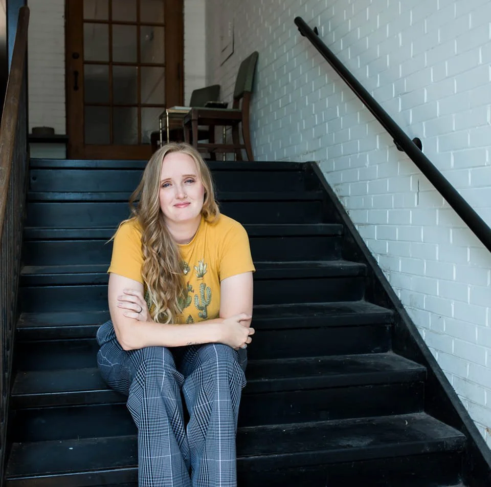 A young woman with long blonde hair and a yellow t-shirt sitting on black wooden stairs in an indoor setting with white brick walls, a metal handrail, and chairs in the background.