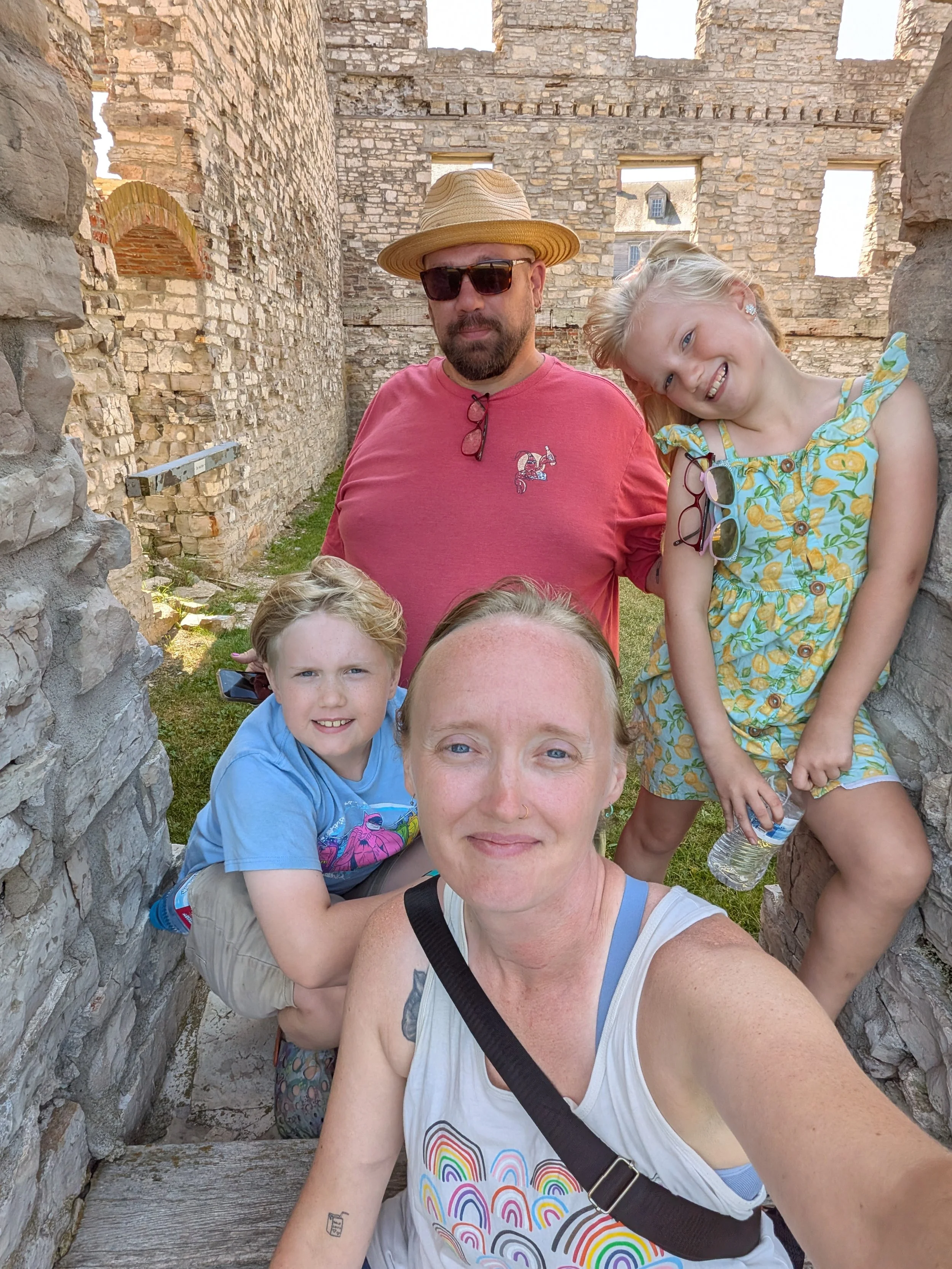 A woman wearing a white shirt is pictured center and behind her are two children smiling and a man wearing a straw hat near a stone structure
