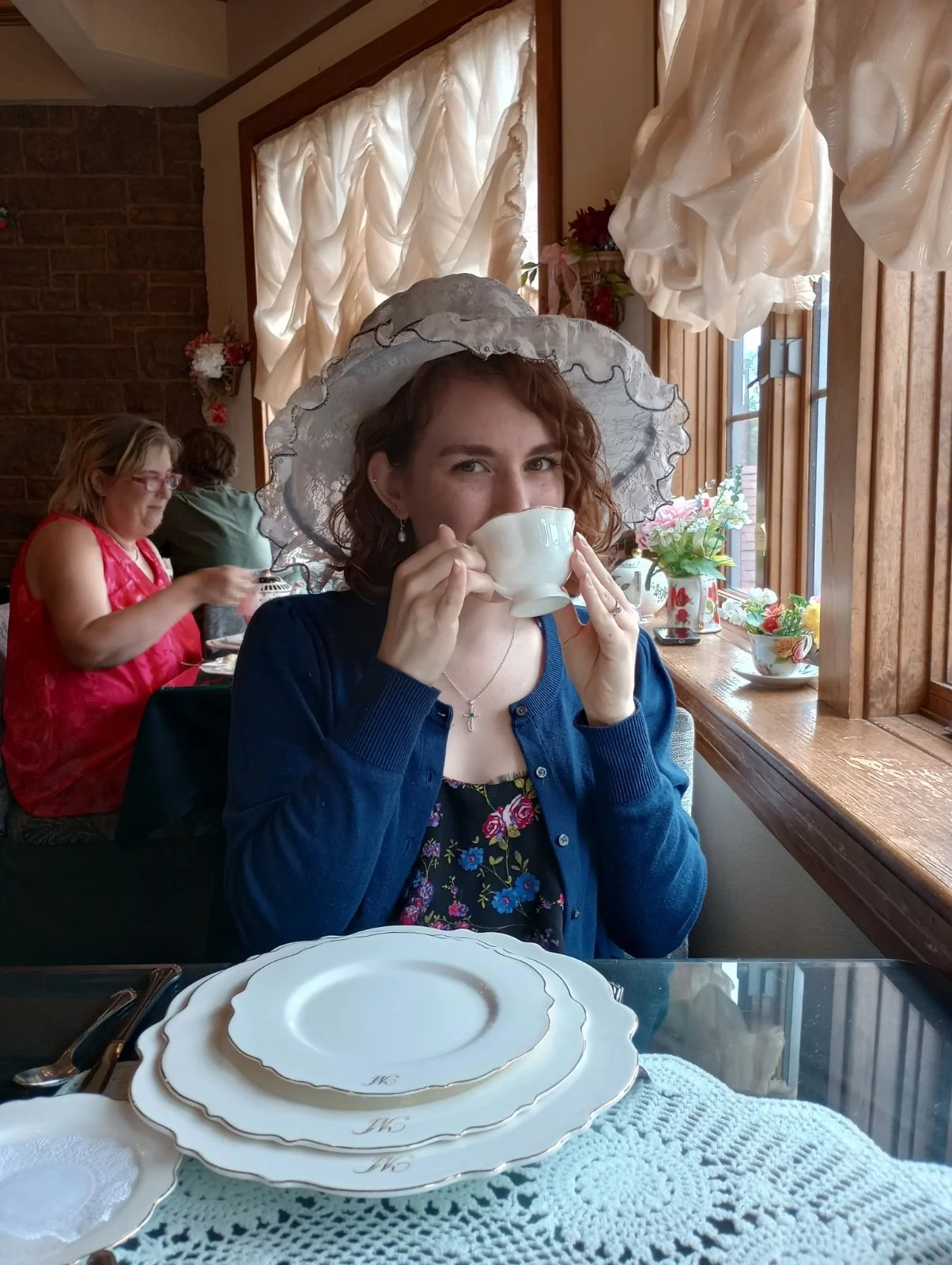 A woman sitting in a tea shop, holding a tea cup, and wearing a white hat