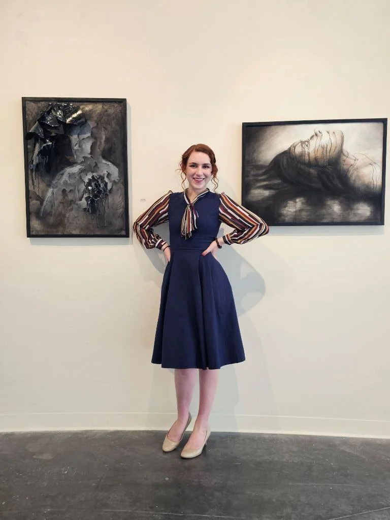 A woman with red hair wearing a navy blue dress with striped long sleeves and a bow tie stands in an art gallery, smiling with her hands on her hips, with two black and white abstract paintings behind her.