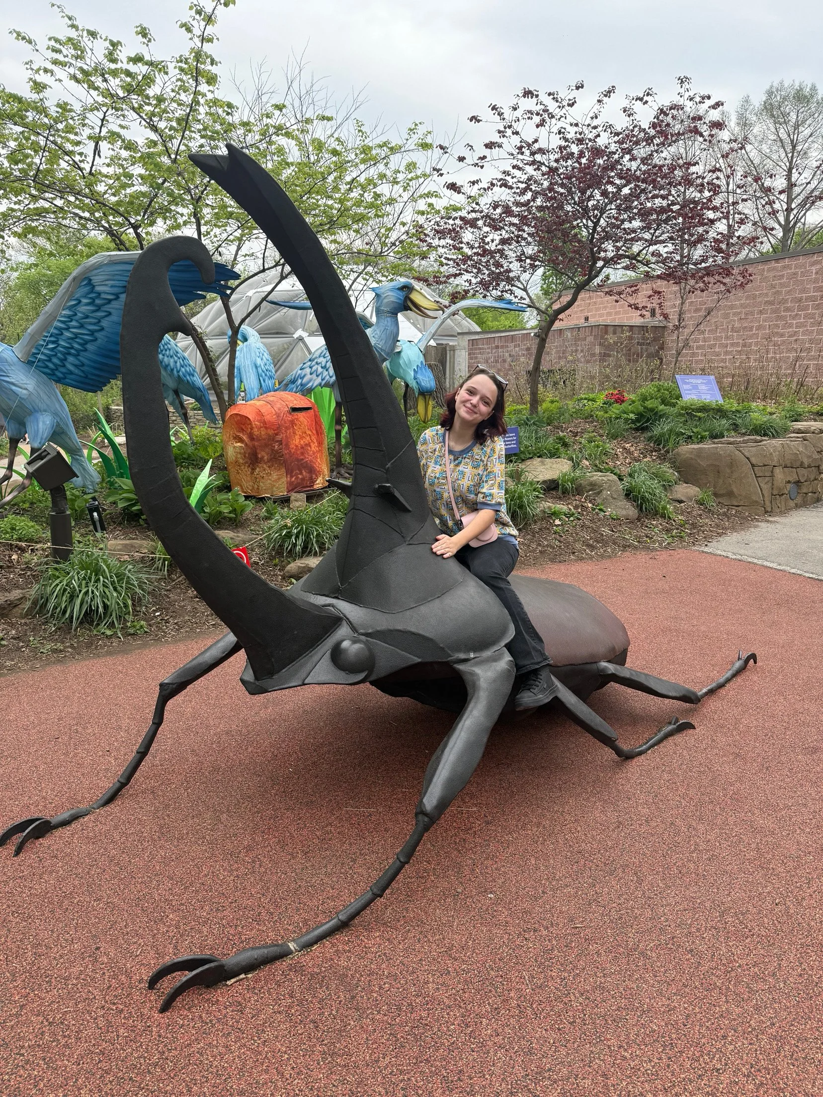 A woman sitting on a large metal sculpture of a black insect with a long body, six legs, and antennae, in a garden with trees and plants.