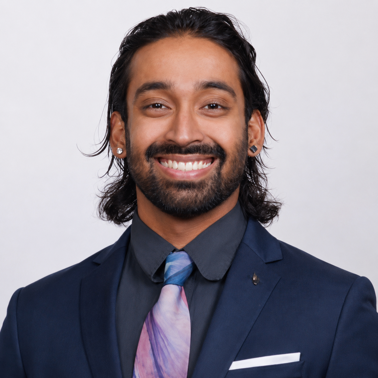 Portrait of a smiling man with dark, wavy hair, wearing a dark suit, black shirt, and colorful tie, against a plain white background.